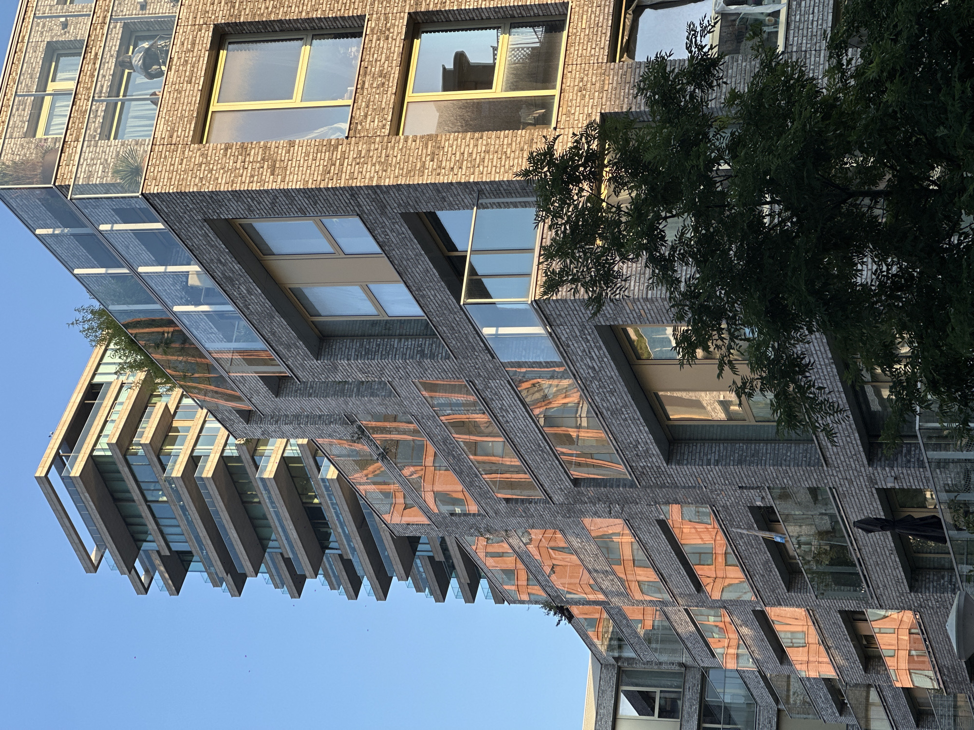 This is a close-up, eye-level, outdoor, daytime photograph of the exterior of a modern apartment building. The building is made of light brown brick and has multiple stories with balconies. The balconies have glass railings. The windows of the building are large and reflect the surrounding environment. The building is modern with clean lines and a contemporary design. In the background, another taller building with multiple stories and balconies is visible. The sky is clear and blue. A tree is in the foreground, partially obscuring the lower part of the building. The image appears to be taken in an urban environment, possibly a city. The question asks for the location in Amsterdam, Netherlands. Given the architectural style of the buildings, this could be a contemporary development in Amsterdam. The reflection on the windows shows an orange colored building, suggesting there is another building of that color nearby.