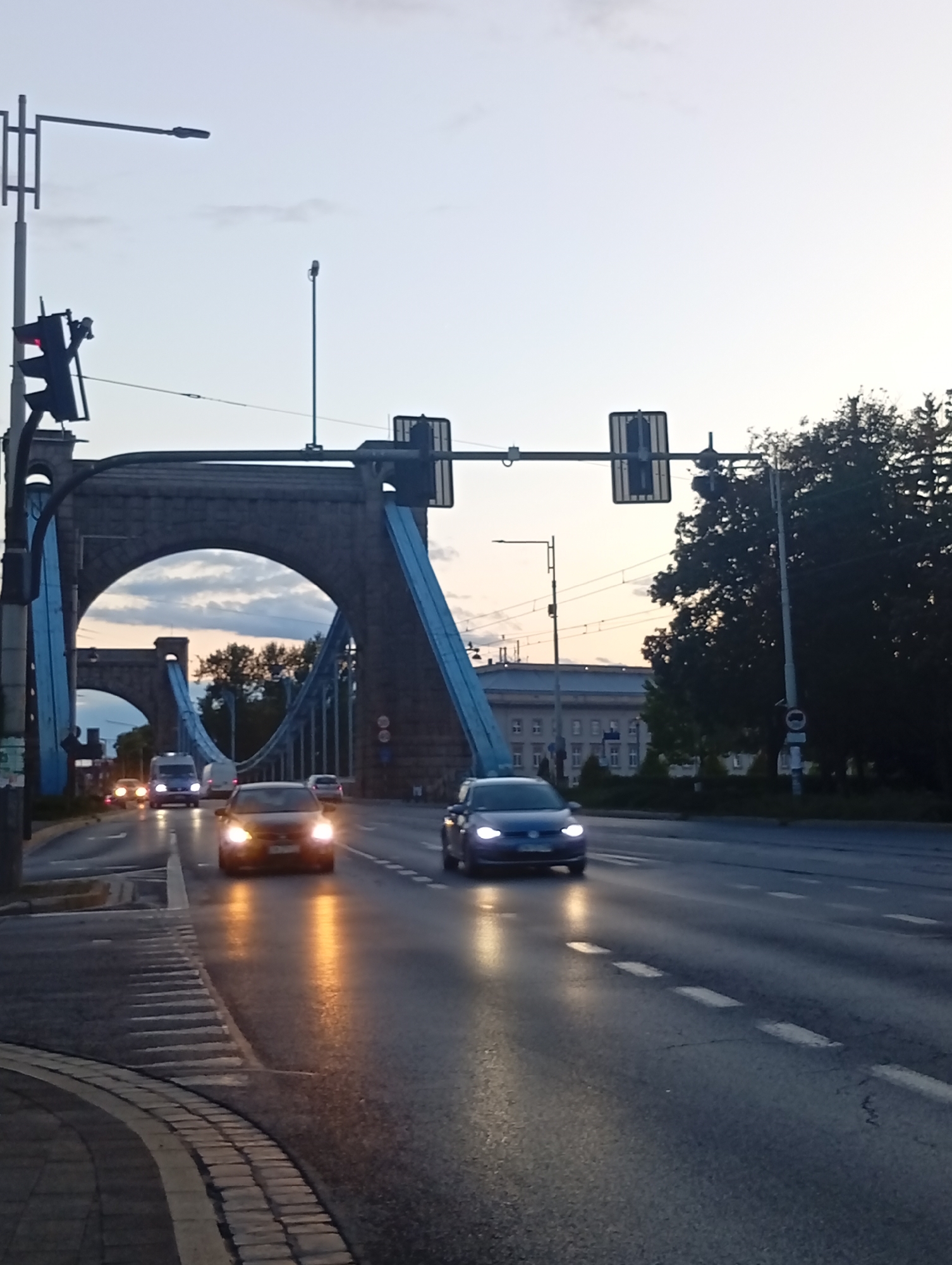 The image depicts a street scene at dusk or dawn, likely on the Most Grunwaldzki (Grunwald Bridge) in Wrocław, Poland. The bridge's distinctive blue suspension elements and large arched stone towers are visible in the background. Cars are driving on the road, with headlights illuminated. One car in the foreground is a dark sedan, possibly a Volkswagen Golf, with its headlights on. Another car, a blue hatchback, is also visible. A white van is seen further back. Traffic lights are positioned overhead, with one showing a red signal. Trees and buildings are visible on the right side of the image, and the sky is a pale blue with some clouds. The road surface is wet, suggesting recent rain. The overall impression is of an urban environment at a transitional time of day.