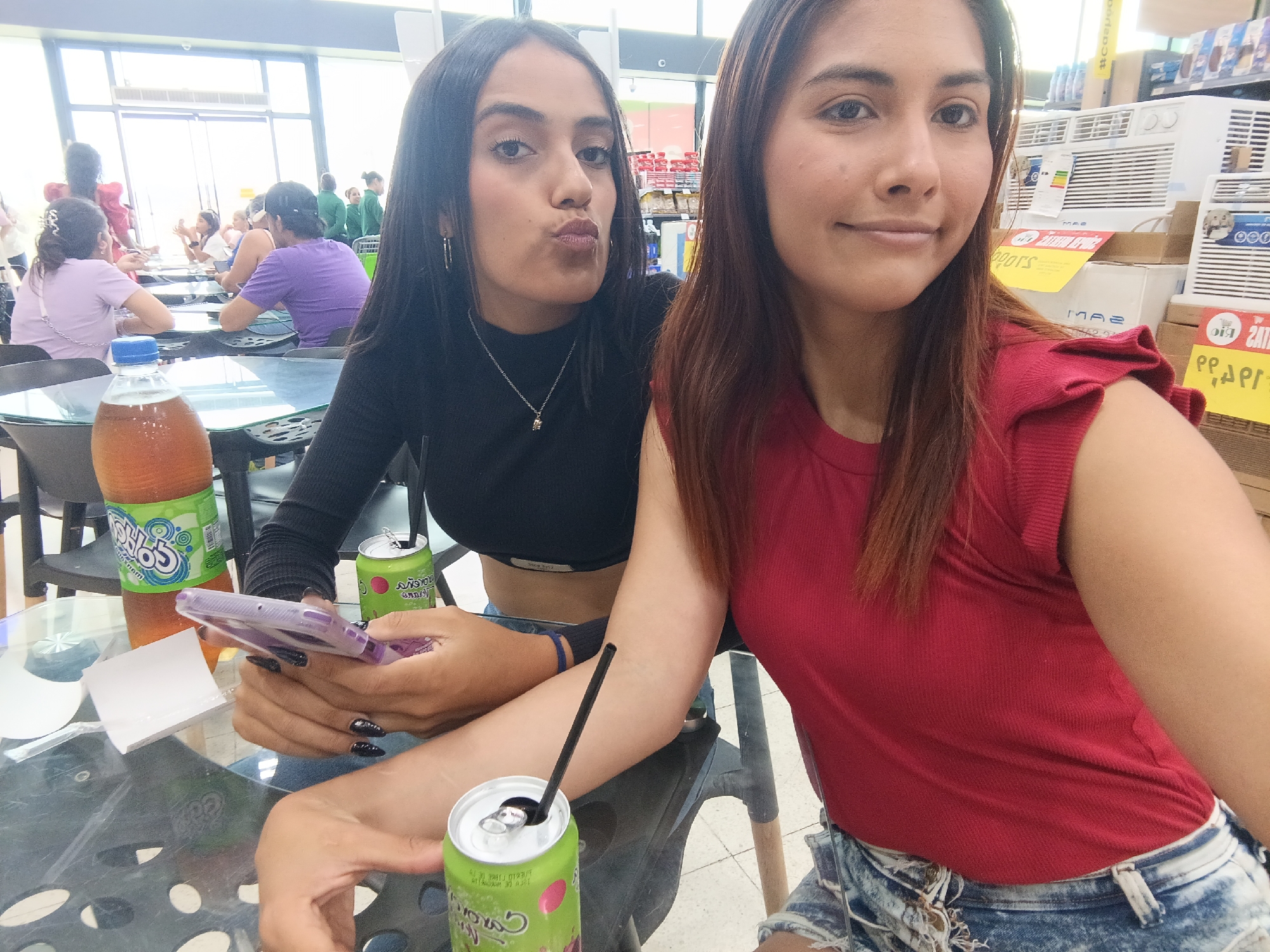 Two young women pose for a selfie in what appears to be a food court or dining area of a store, possibly in Juan Griego, Venezuela, given the reference to that location. The woman on the left wears a black long-sleeved crop top and jeans and is holding a smartphone. Her lips are pursed in a kissy face. She has long dark hair and is wearing a silver necklace. The woman on the right wears a red shirt and acid-wash denim shorts. She has medium-length brown hair and is smiling. On the table are drinks including a large bottle of "Chilos" brand soda and cans of "Sangria Caroreña" soda with one can having a straw. In the background are other people seated at tables, and shelves of products, including what appears to be air conditioners. The lighting suggests a bright, possibly indoor environment.