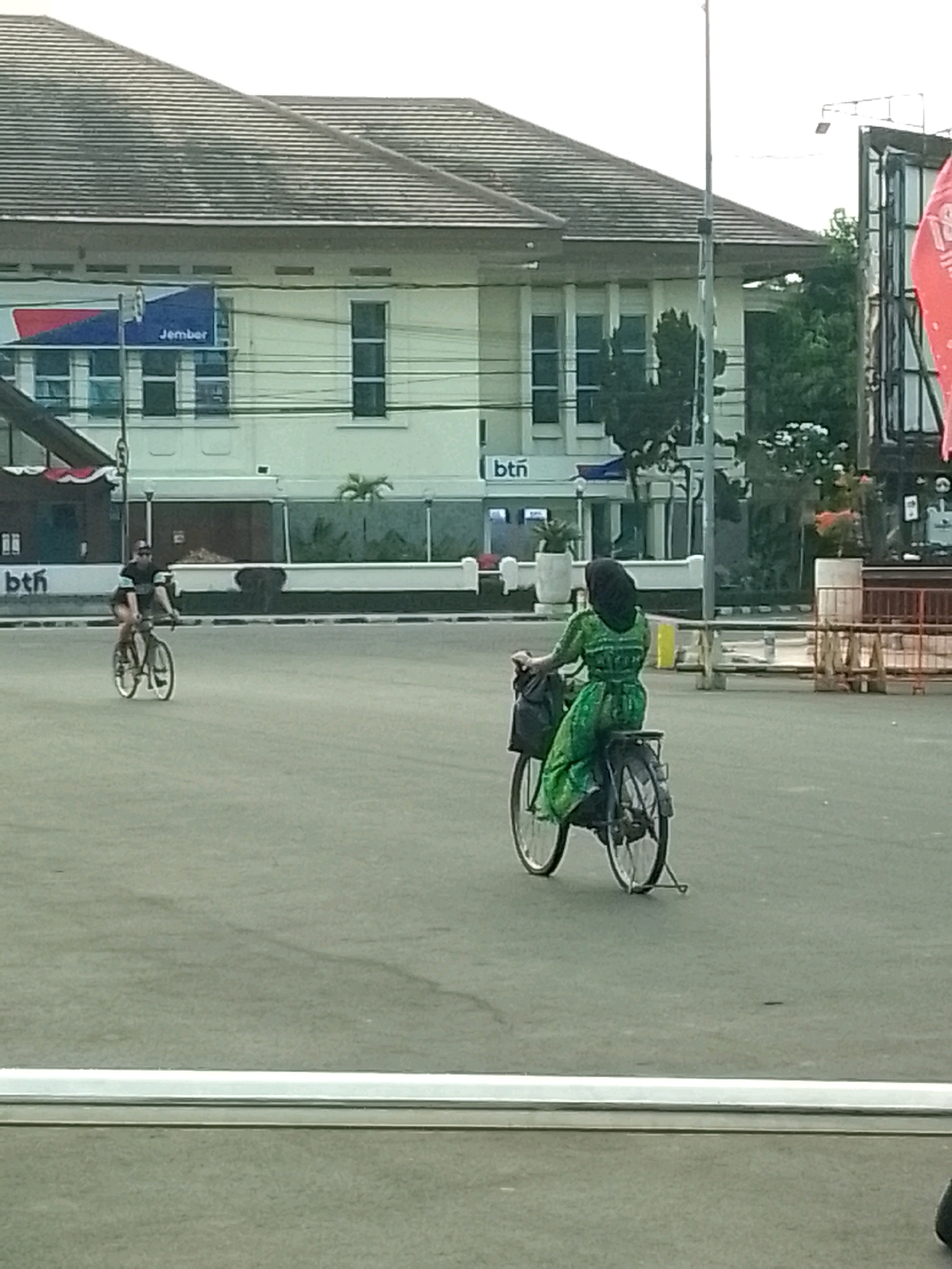 In Jember, Indonesia, an outdoor, urban scene unfolds. The focal point is a woman on a bicycle, her back to the camera, wearing a green patterned dress and a black hijab. Her bicycle has a black bag attached to the rear rack. Another cyclist, a person in a black shirt and shorts, rides a bicycle in the background, to the left. They are on a paved road, with a white curb and a white line marking the edge of the road. Behind them stands a building with a tiled roof and windows. A sign on the building reads "Jember". Further back, trees and a red banner are visible.