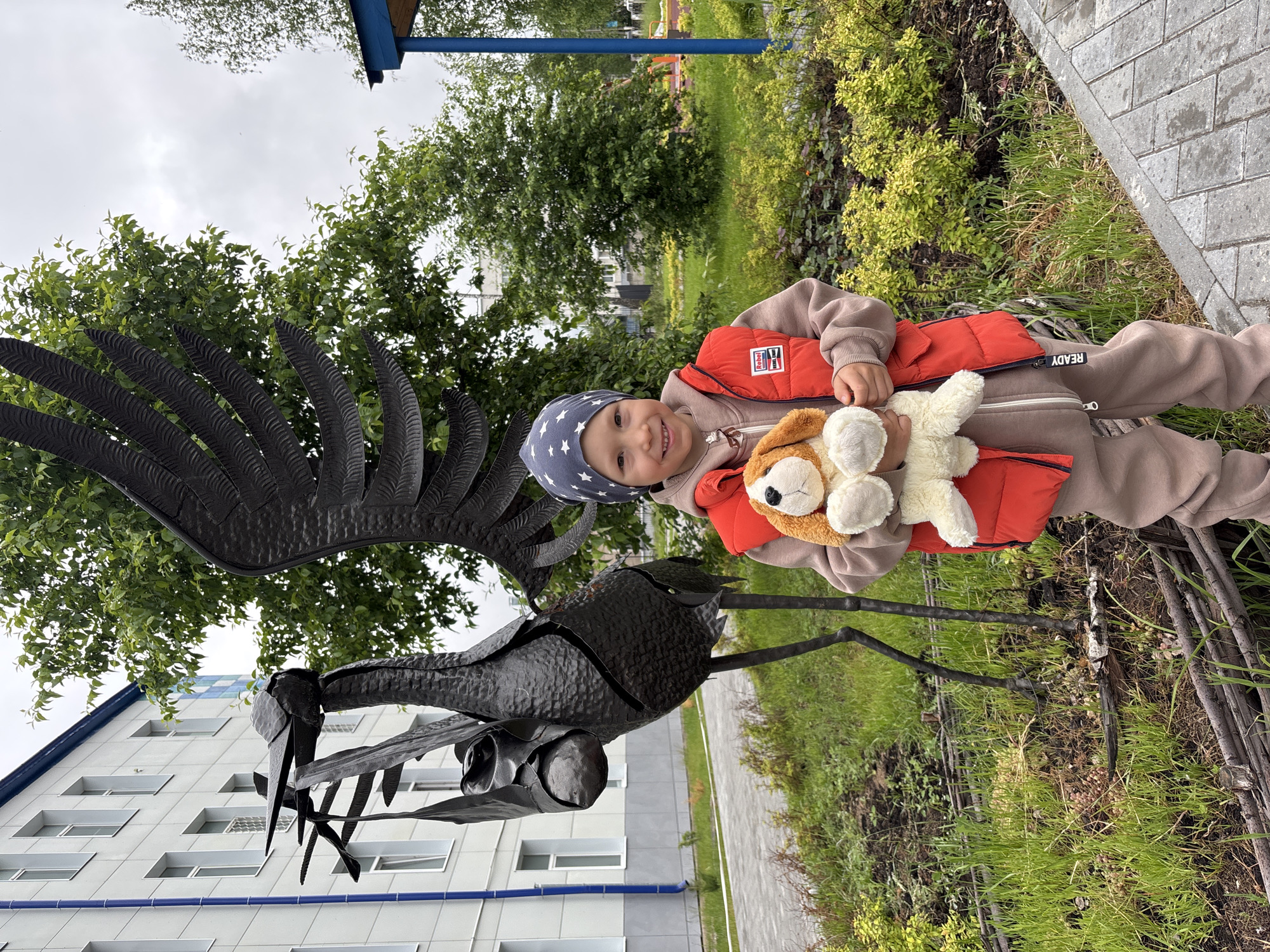 This image, captured in Chaikovsky, Russia, presents a young child standing outdoors next to a large metal sculpture. The child, a boy with light skin, is positioned slightly off-center to the right, smiling directly at the viewer. He wears a blue beanie with white stars, an orange quilted vest over a beige hooded tracksuit, and beige pants with "READY" written vertically on the left leg. He holds a light brown and white stuffed dog in both hands at waist level.
To the child's left and extending upward, a tall, dark metal sculpture dominates the left side of the frame. It depicts a stylized bird, resembling a stork or crane, with large, textured wings splayed to the left and rear. The bird's head and long beak are pointed slightly upwards and to the left, and it appears to be carrying a wrapped bundle or object suspended from its beak area. The sculpture stands on thin, dark legs that are embedded in a patch of green grass and exposed soil.
In the background, a multi-story building with light-colored (likely white or light grey) walls and numerous rectangular windows is visible on the left. A vertical blue drainage pipe runs down the left side of the building. The upper edge of the building shows a distinct blue roofline. Behind the child and the sculpture, a lush green environment features various trees with green foliage, suggesting an outdoor park or landscaped area. A blue vertical support beam, possibly part of a canopy or small structure, is visible on the far right. A paved walkway made of grey pavers is present in the lower right foreground, bordering the grassy area. A low, rustic fence constructed from interwoven branches separates the paved path from the garden bed in the lower center. The sky is overcast and light grey.