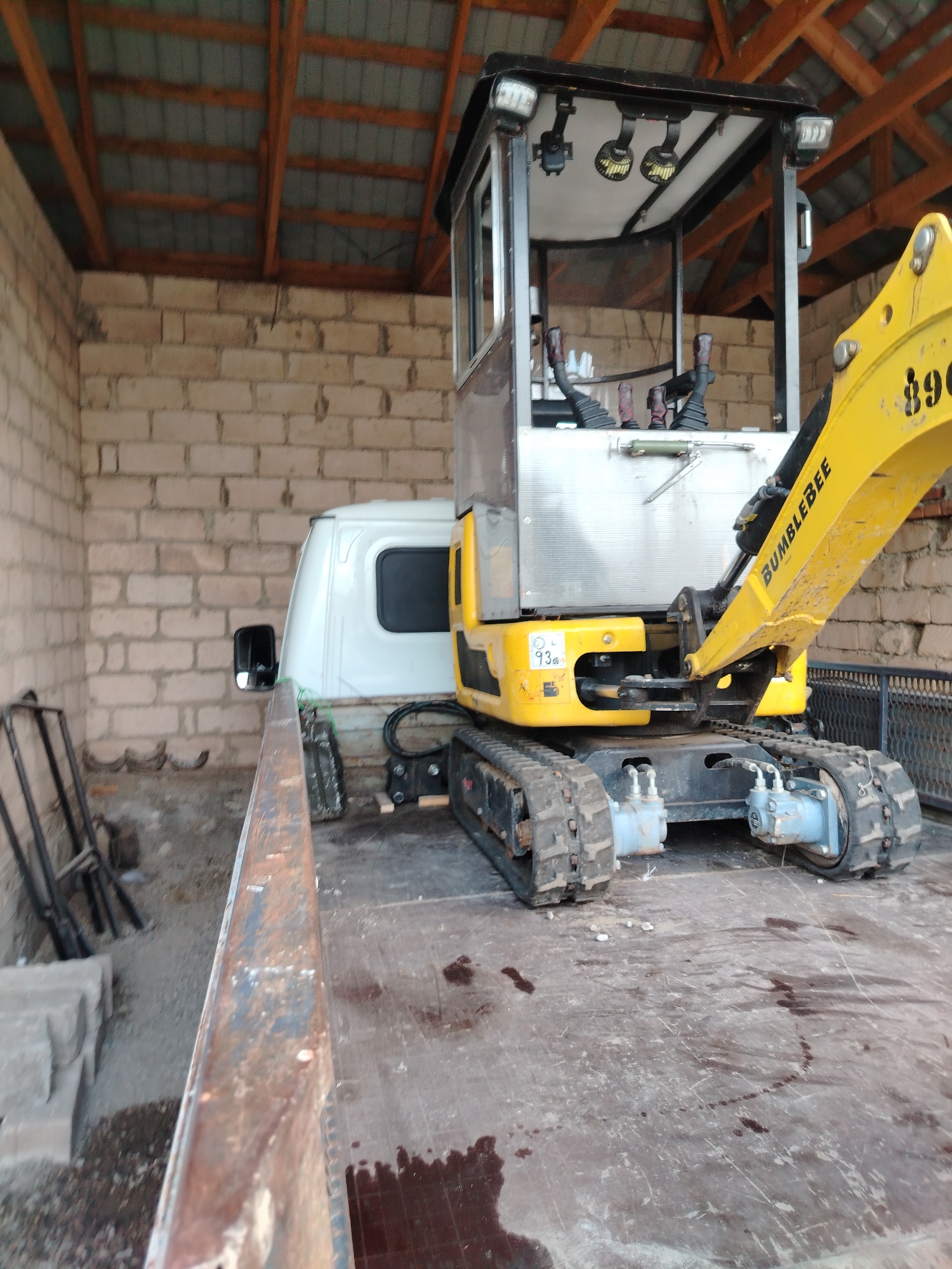A yellow and black compact excavator, identified by the text "BUMBLEBEE" on its arm, is parked on the flatbed of a white truck. The excavator has a closed cab with windows, two lights on the roof, and control levers inside. Its tracks are visible, as are hydraulic components. The truck is positioned inside a rustic, open-sided structure with a wooden-beamed roof and cinder block walls. The ground is covered in gravel and dirt, with some debris. The overall scene suggests a storage or workshop area in Атажукино, Russia.