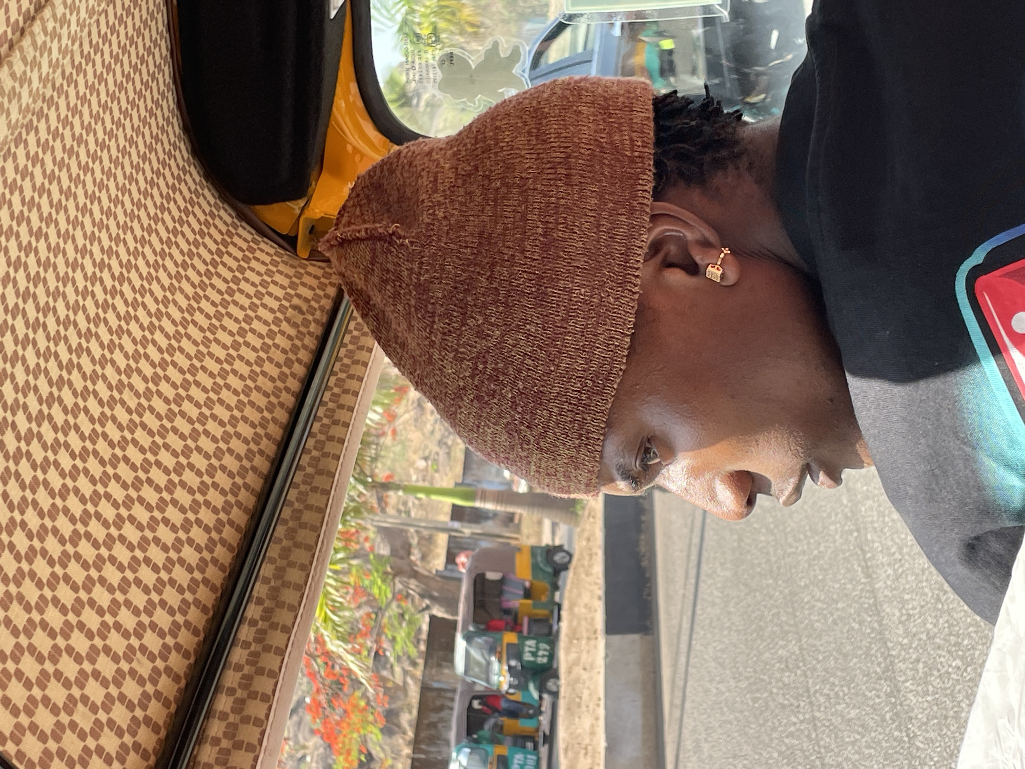 A close-up shot presents a person with dark skin and short dark hair, wearing a brown knit hat and a black t-shirt with a colorful graphic design. The person's right ear is visible, adorned with a gold earring. They are inside a vehicle with patterned upholstery, likely an auto rickshaw, also known as a "Keke Napep." Through the window, the outdoor environment reveals a road lined with trees, red flowers, and parked Keke Napeps. The scene indicates a location in Jos, Nigeria.