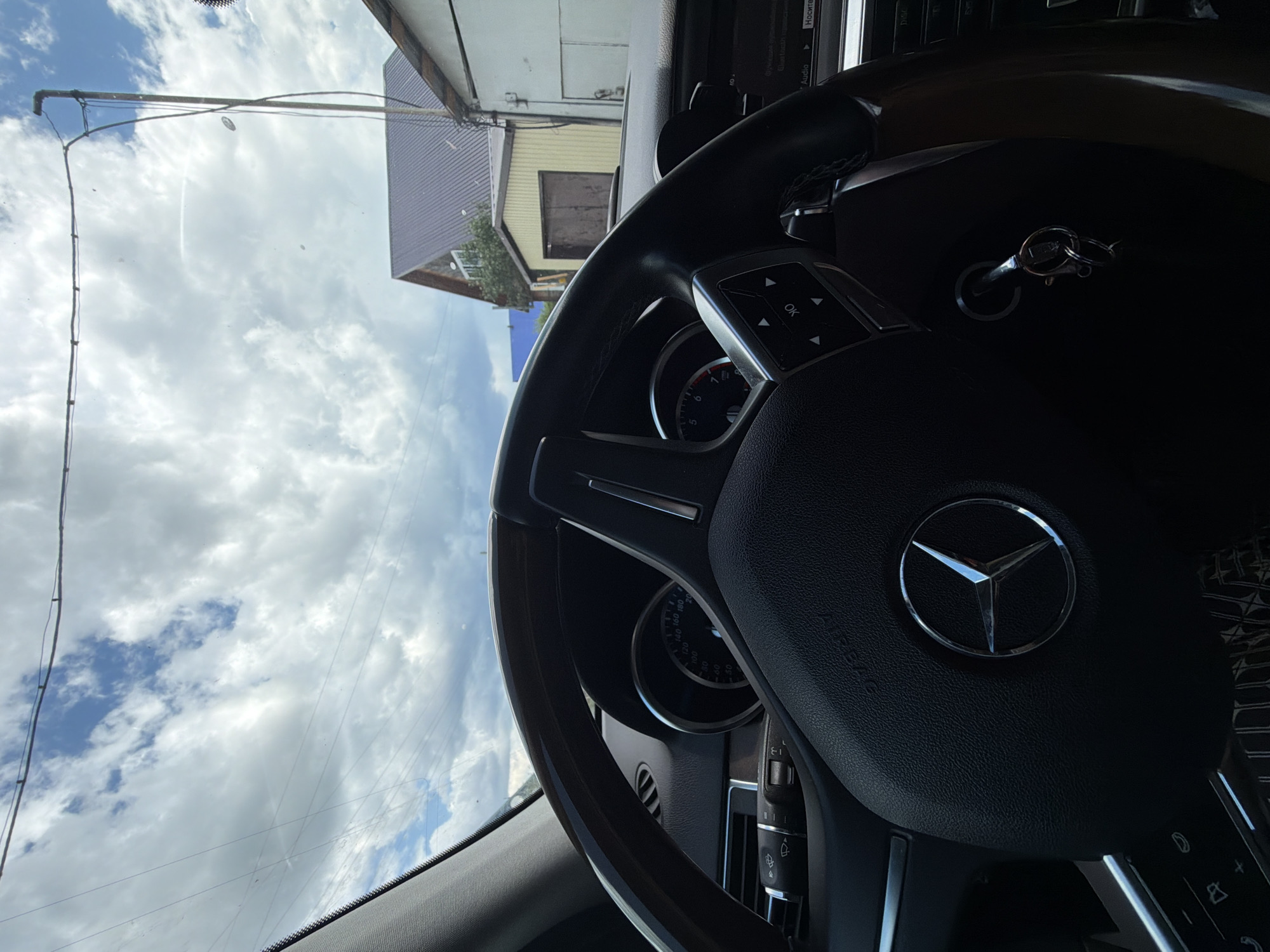 The image is a close-up interior shot of a Mercedes-Benz car, focusing on the steering wheel and dashboard. The car is parked outdoors, with a view of a building and a cloudy sky through the windshield. The steering wheel is black leather with wood trim and features the Mercedes-Benz logo in the center. A car key is inserted into the ignition. The dashboard shows a speedometer and other instrument clusters. In the background, a multi-story building with a dark, sloped roof is visible, as well as a smaller yellow structure with a garage door. The sky is filled with white and gray clouds. The scene appears to be in a residential or semi-rural area. The location is the city of Mezhevoy, Russia.