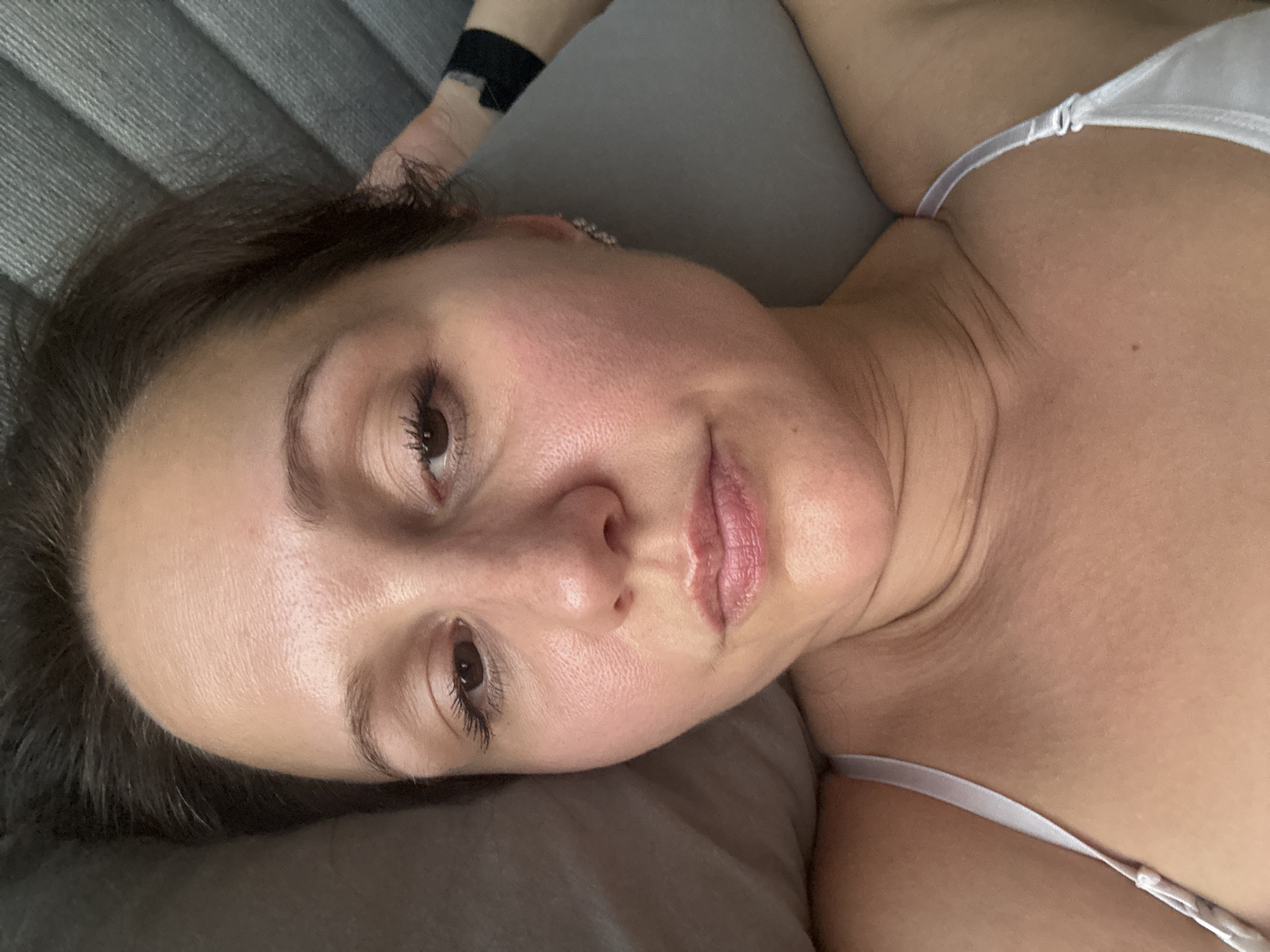A woman is lying down, her face close to the camera. She has brown hair, brown eyes, and appears to be wearing a white top. Her skin has a reddish tone. The background consists of a grey cushion and what appears to be a headboard. A black object, possibly a wristband, is visible on her left arm. The image does not appear to be taken in Dubai, United Arab Emirates.