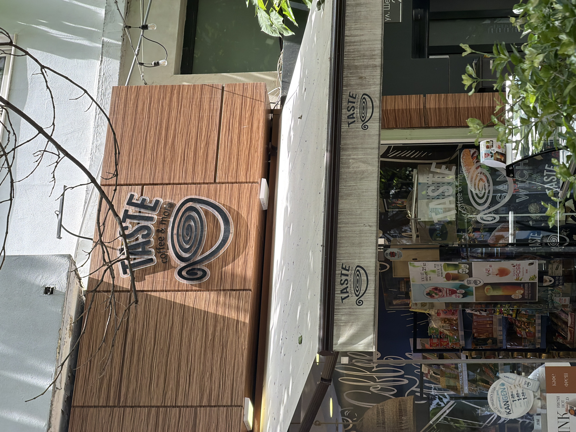 The image shows the exterior of a coffee shop called "TASTE coffee & more" located in Varna, Bulgaria. The shop has a light-colored awning with the "TASTE" logo. Above the awning, a larger sign with the "TASTE coffee & more" logo is mounted on a wooden-paneled wall. The interior of the shop is visible through the glass windows, displaying shelves stocked with products and various promotional posters. In the foreground, there are green plants and branches from a tree are seen in the upper portion of the image. The lighting suggests it is daytime, with shadows cast on the building.