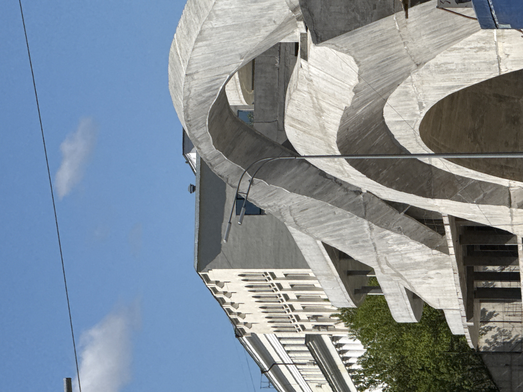 The image depicts the exterior architecture of the MÖİ Stadium in Kazan, Russia. The architecture includes a series of curving concrete elements. These curves surround a more standard building with windows. The building and surrounding landscape are shot from a low angle. The sky above is clear and blue. A thin power line crosses the image on the left side.