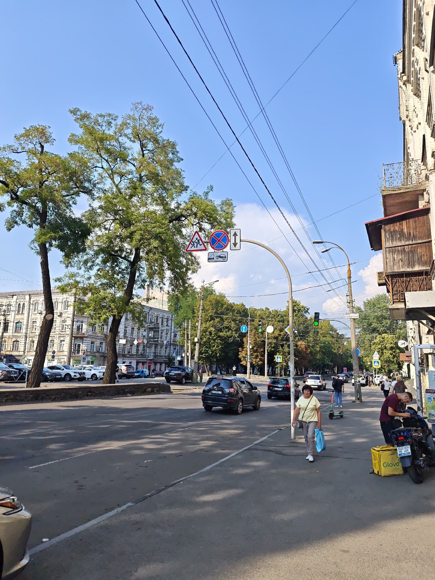 The image shows a street scene in Kyiv, Ukraine, on a sunny day with a clear blue sky. The street is lined with trees and multi-story buildings. Several cars are parked along the side of the road and others are driving on the street. Pedestrians are visible on the sidewalk. One woman is walking with a blue shopping bag, and another person is standing next to a motorcycle with a yellow delivery bag. There are traffic lights and road signs indicating traffic regulations. Overhead power lines are strung across the sky. The overall impression is of a bustling urban environment.