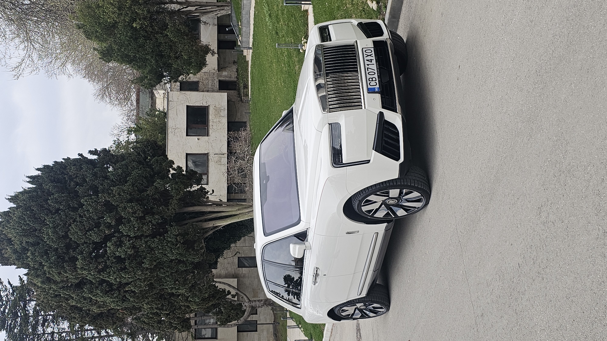 A white Rolls-Royce Cullinan is parked on an asphalt road. The license plate reads "CB 0714 XO" and indicates Bulgarian registration. The car is positioned next to a building with multiple windows and adjacent to greenery, including trees and grass. Based on the architectural style and vehicle registration, the location is likely in Varna, Bulgaria. No people are visible in the image. The overall scene suggests a residential or suburban area.