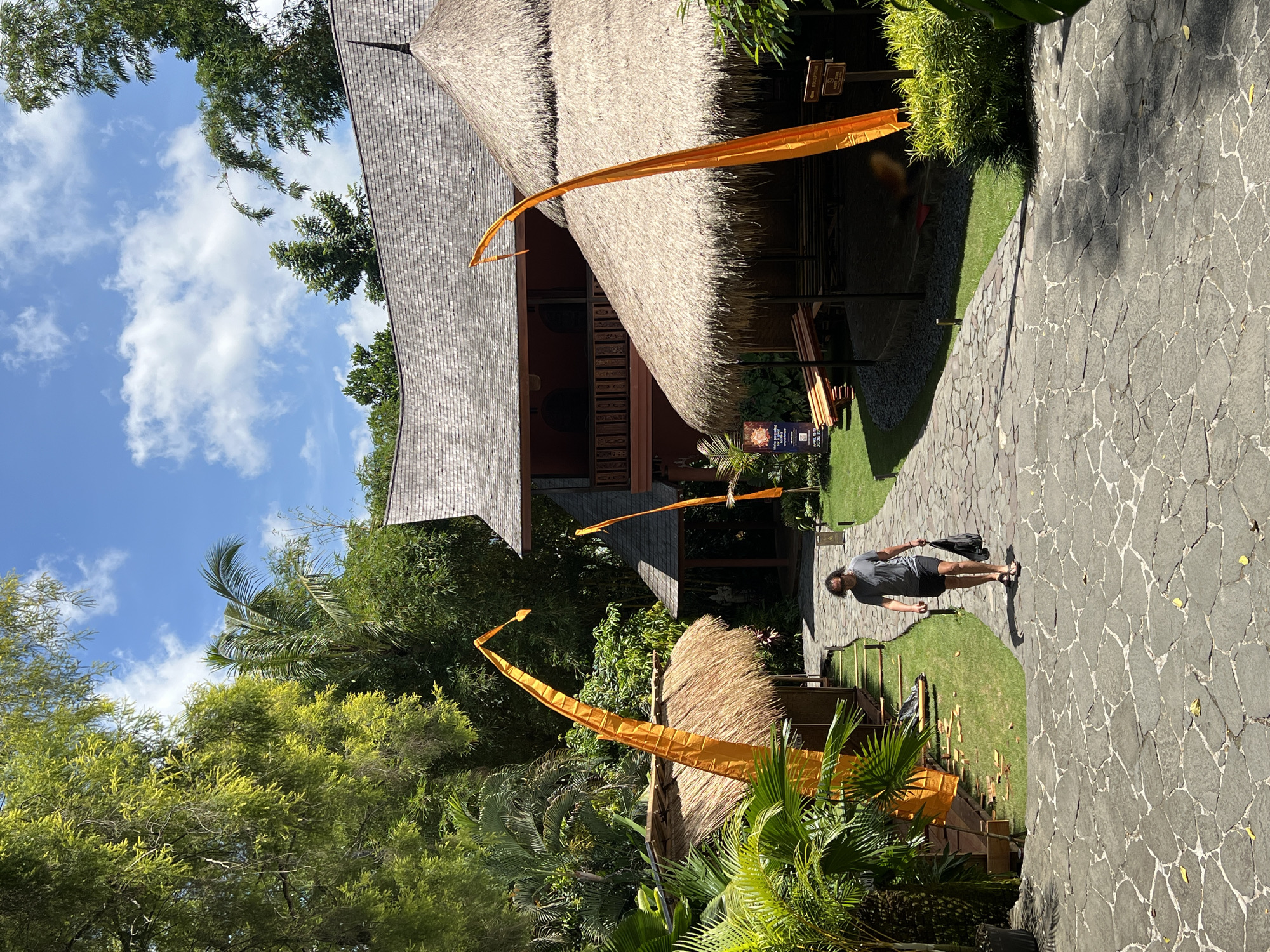 The image depicts a man walking on a stone pathway in Peliatan, Indonesia. The path leads past structures with thatched roofs and distinctive orange flags. Lush greenery surrounds the buildings. The sky above shows scattered clouds.