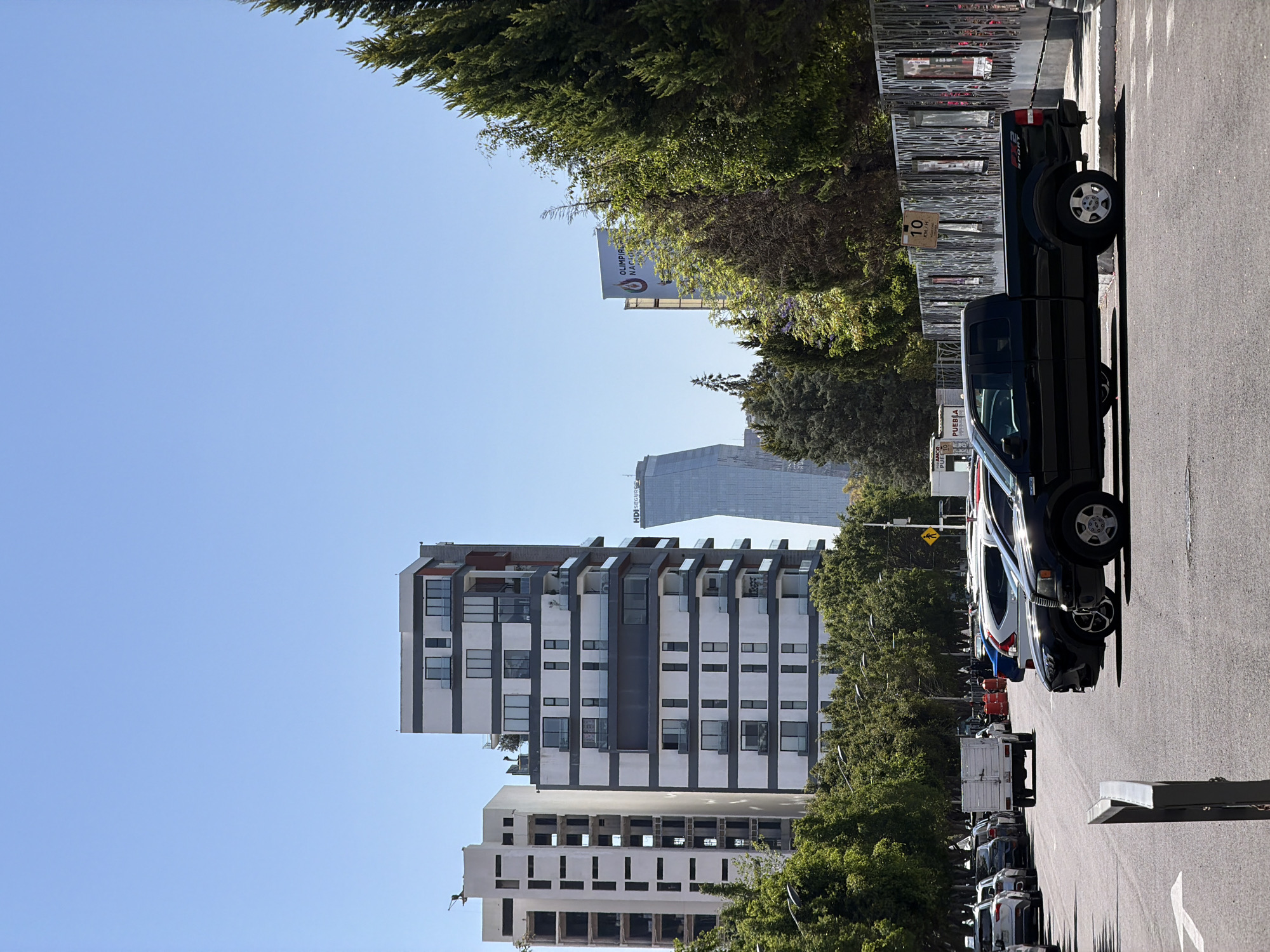 The image, rotated 90 degrees clockwise, depicts a sunny urban street scene. If viewed upright, a clear, light blue sky would dominate the upper portion of the frame, with a road at the bottom and buildings and trees lining the sides.
In the midground, a modern apartment building with white and dark gray sections features numerous balconies, some with glass railings. Below it, another building with a lighter, perhaps unfinished, facade and multiple rectangular openings is visible. In the background, a tall, sleek, glass-fronted skyscraper, marked with "HDI SEGUR S" (likely "HDI SEGUROS"), rises above other structures.
Lush green trees are abundant, lining the street and partially obscuring a large billboard. The asphalt road on the right side of the image (bottom if upright) shows white lane markings. Several vehicles are parked along the curb, facing the viewer. A prominent black pickup truck, possibly a Ford F-150 with "SP RT" (likely 'SPORT') visible on its side, is in the foreground, featuring a spare tire mounted on its bed. Other cars and a white truck/van are parked further down the street. A decorative metal fence with horizontal slats runs alongside the road, behind the black pickup truck.
Visible text includes "OLIMPIAD NACIO AL" (part of "OLIMPIADA NACIONAL") and a related logo on the billboard partially hidden by trees. A street sign behind the black pickup truck displays "10," possibly a speed limit. Text "PUEBLA" and "PI...ES" can be seen on a white vehicle further down the street.
The scene suggests a calm, bright day, likely morning or early afternoon, in an urban setting within Puebla, Mexico. There are no people visible, and the overall atmosphere is one of a quiet, established city street.
