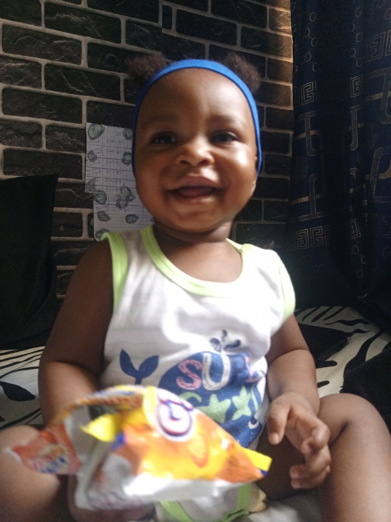 A young baby or toddler is pictured sitting indoors, smiling brightly at the camera. The child has dark skin and is wearing a blue headband with small ear-like protrusions, and a white sleeveless top with a blue and green graphic design featuring a whale-like tail, star shapes, and partial letters like "SU" or "SUI". In their hands, the baby holds a colorful, partially opened snack bag, appearing to be orange, yellow, red, and blue, possibly depicting a cartoon fish or animal.
The background features a dark, textured wall with a brick pattern on the left, and a dark curtain with repetitive patterned letters, including "G" and "C" (reminiscent of a luxury brand logo), on the right. Taped to the brick wall is a white sheet of paper with handwritten text, appearing to be a schedule or list. Visible text includes "ENG 411", "ENG412", "ENG 413", "ENG 414", and "LENGAN", along with a date "07/18/2023". A black cushion or pillow is visible on the far left, and a black and white striped sheet or cushion is beneath the child. The overall scene suggests a casual, happy moment within a home environment during daytime.