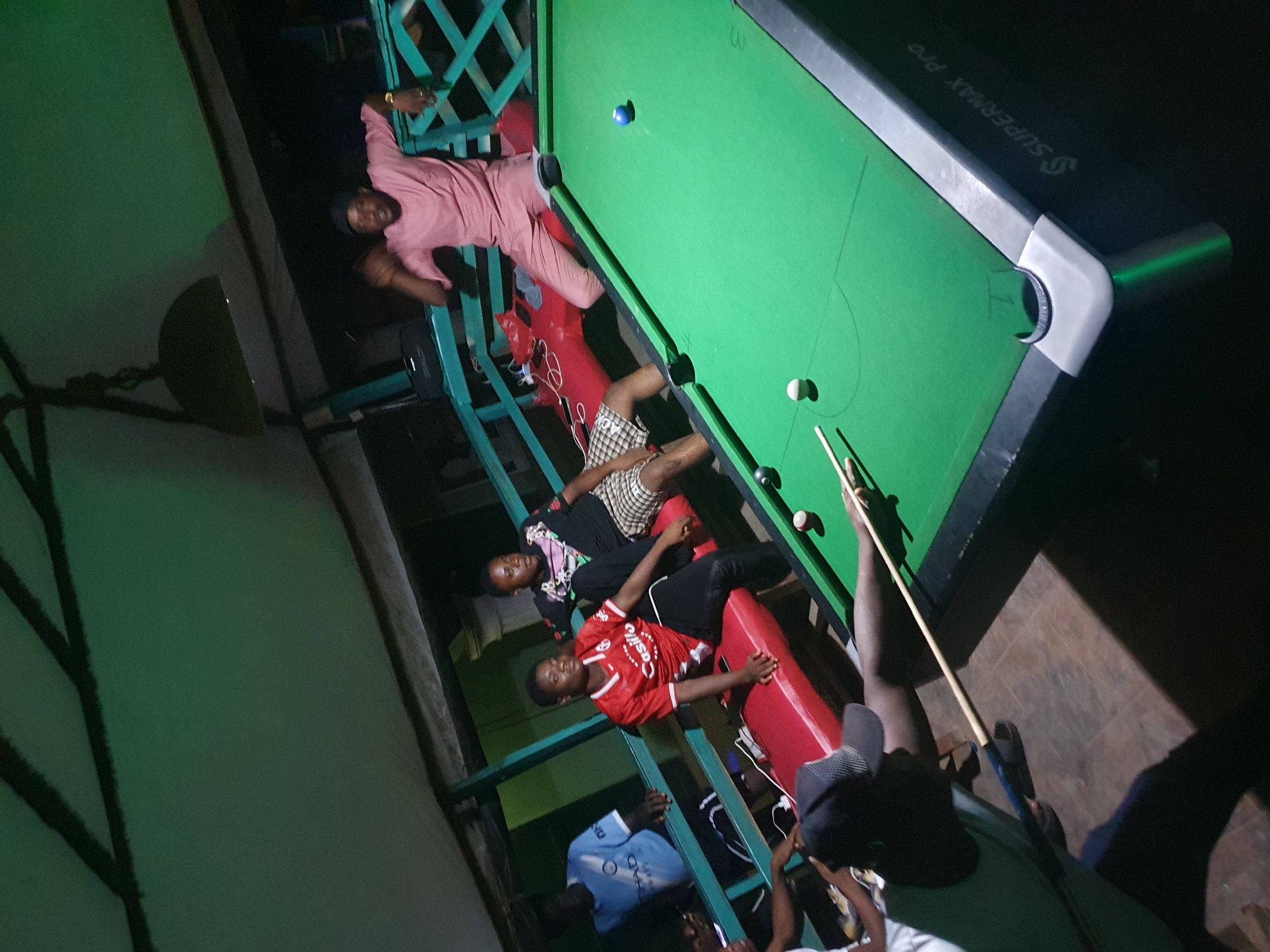 A nighttime scene shows a group of young men in Lokoja, Nigeria, around a pool table. One man, wearing a pink outfit and cap, is reclining on a teal green metal bar surrounding the table. Below him, two men sit on a red padded area with black metal trim, one in a black patterned shirt and the other in a red jersey with a white logo. One man is taking a shot with a cue stick. Other individuals are visible below the teal bar. The pool table is green with a gray and white border, marked "Supermax Pro." Pool balls are scattered on the table. A dim light casts shadows in the background.