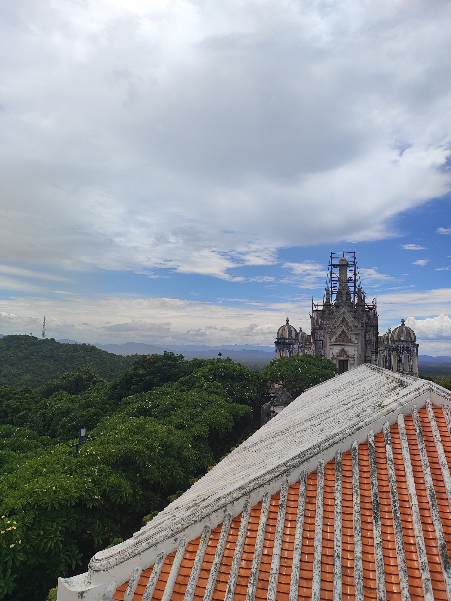This is an outdoor, high-angle shot of the Maruekhathaiyawan Palace in Phetchaburi, Thailand, on a cloudy day. In the foreground, the tiled roof of a building, made of orange tiles and white concrete, occupies the lower right portion of the frame. Beyond the roof, lush green trees with scattered white flowers stretch across the middle ground, leading to rolling hills in the background. A telecommunications tower is visible on a distant hill. In the middle right of the image, the ornate, multi-domed structure of the Maruekhathaiyawan Palace is partially obscured by scaffolding, suggesting it is undergoing renovation. The sky is filled with a mix of white and gray clouds, with patches of blue sky visible.