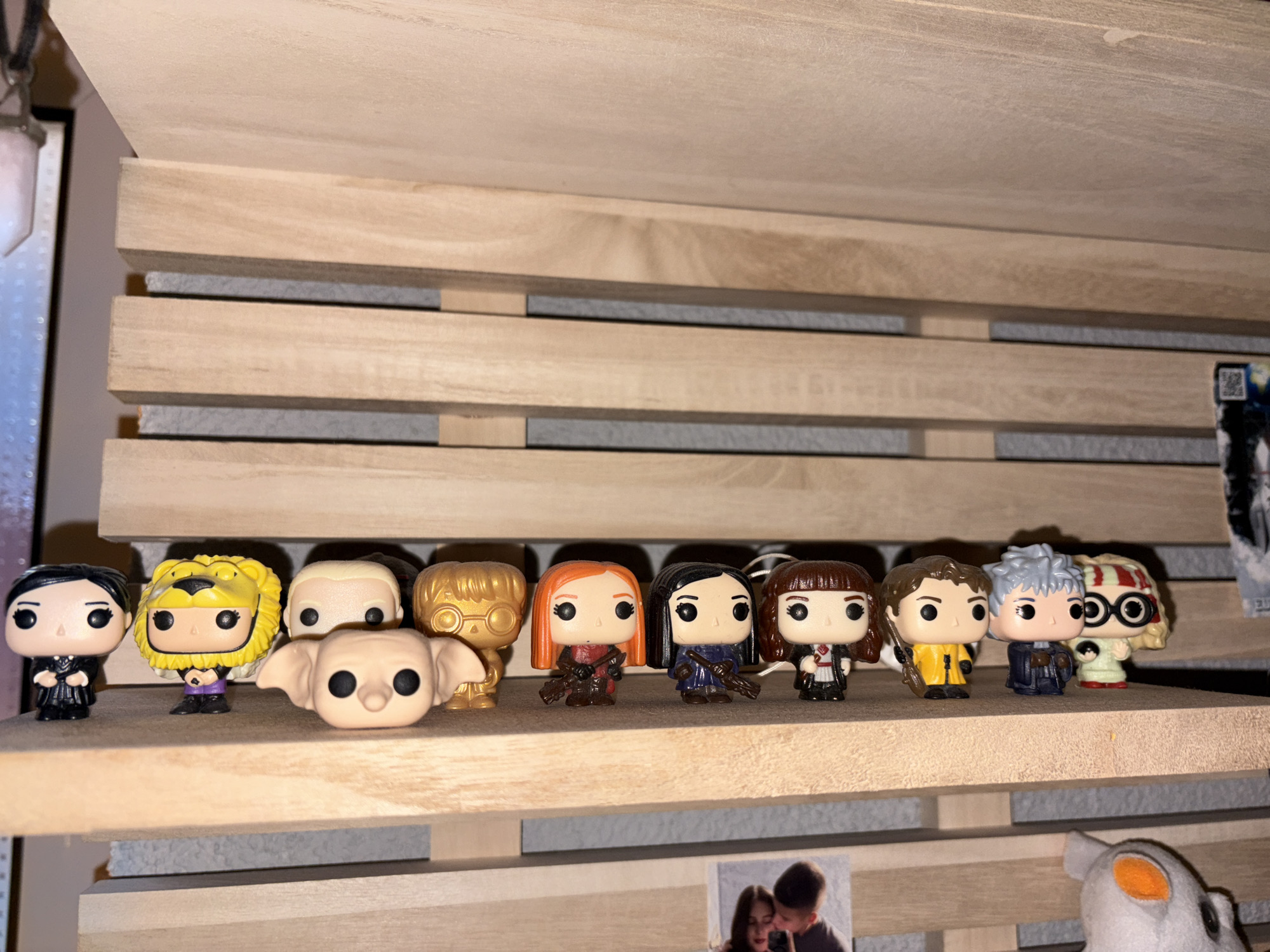 The image shows a collection of Funko Pop figurines displayed on a light-colored wooden shelf with horizontal slats. The figurines appear to be Harry Potter characters. From left to right, the figures include: an individual in a black dress; an individual in a yellow lion costume; Dobby the House Elf; a blank figure; golden Harry Potter; Ginny Weasley; Bellatrix Lestrange; Hermione Granger; Ron Weasley; Remus Lupin; and Luna Lovegood. Another shelf is visible below, containing a photo of a couple and a white stuffed toy with an orange eye. The shelf is in a location in the city of Panagyurishte, Bulgaria.