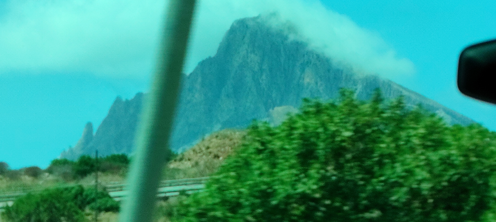 The media file captures a mountainous landscape, likely taken from a moving vehicle due to the slight blur and the presence of a dark object in the upper right corner, possibly a side mirror. The primary subject is a large, rugged mountain that dominates the background, partially obscured by wisps of clouds. The sky is a bright, clear blue, suggesting daytime. In the foreground, lush green foliage is visible, adding a natural element to the scene. The overall impression is one of a scenic drive through a mountainous region. There is no visible text in the image.