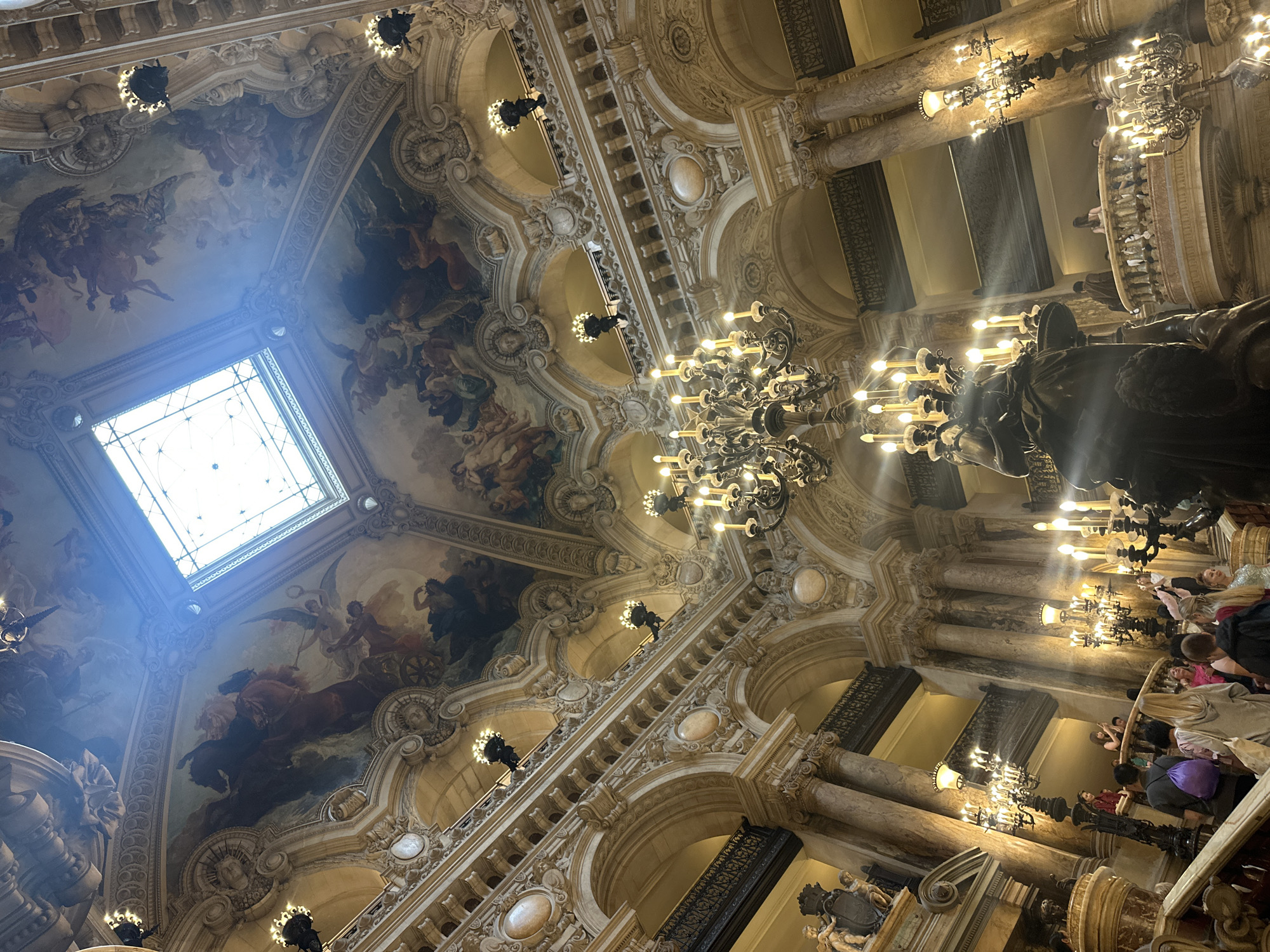 This image captures the opulent interior of a grand, historical building, likely the Palais Garnier (Opéra Garnier) in Paris, France, viewed from a low angle looking upwards. The dominant feature is the elaborately decorated ceiling, which is adorned with intricate frescoes depicting mythological or allegorical scenes featuring figures, winged beings, horses, and chariots. A large, rectangular skylight on the upper left side allows bright natural light to stream in, creating a noticeable lens flare.
The ceiling is supported by rich architectural elements, including ornate moldings, carvings, and coffered sections. Numerous multi-tiered chandeliers, featuring multiple electric candles, hang from the ceiling, casting a warm, artificial glow that complements the natural light.
On the right side of the image, the view extends down towards what appears to be a grand staircase or hall. Marble columns, detailed balustrades, and arches are visible, further emphasizing the grandeur of the setting. Large, dark-colored sculptures or statues are positioned along the architectural features.
Several people, appearing to be tourists or visitors, are visible in the lower right and along the upper levels. They are casually dressed and appear to be observing and admiring the magnificent architecture and artwork. The scene conveys a sense of awe and historical grandeur. The presence of natural light through the skylight indicates it is daytime. There is no visible text in the image.