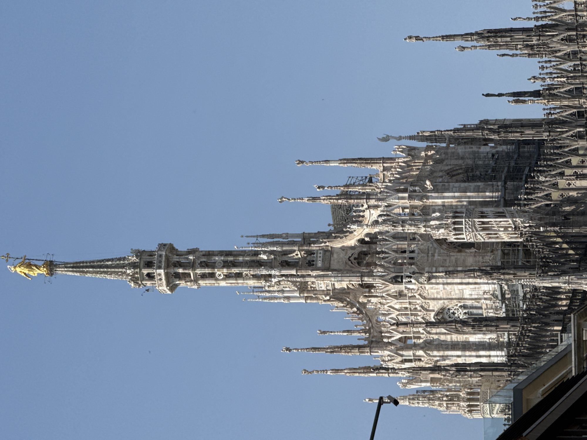 The image depicts the roofline and spires of the Milan Cathedral (Duomo di Milano) in Milan, Italy, against a clear blue sky. The architectural details are highly intricate and ornate, characteristic of Gothic style. At the leftmost edge of the structure is a tower with a gold-colored statue at its pinnacle. In the lower right corner, there is a dark roofline of a structure beneath it, possible a building roof.