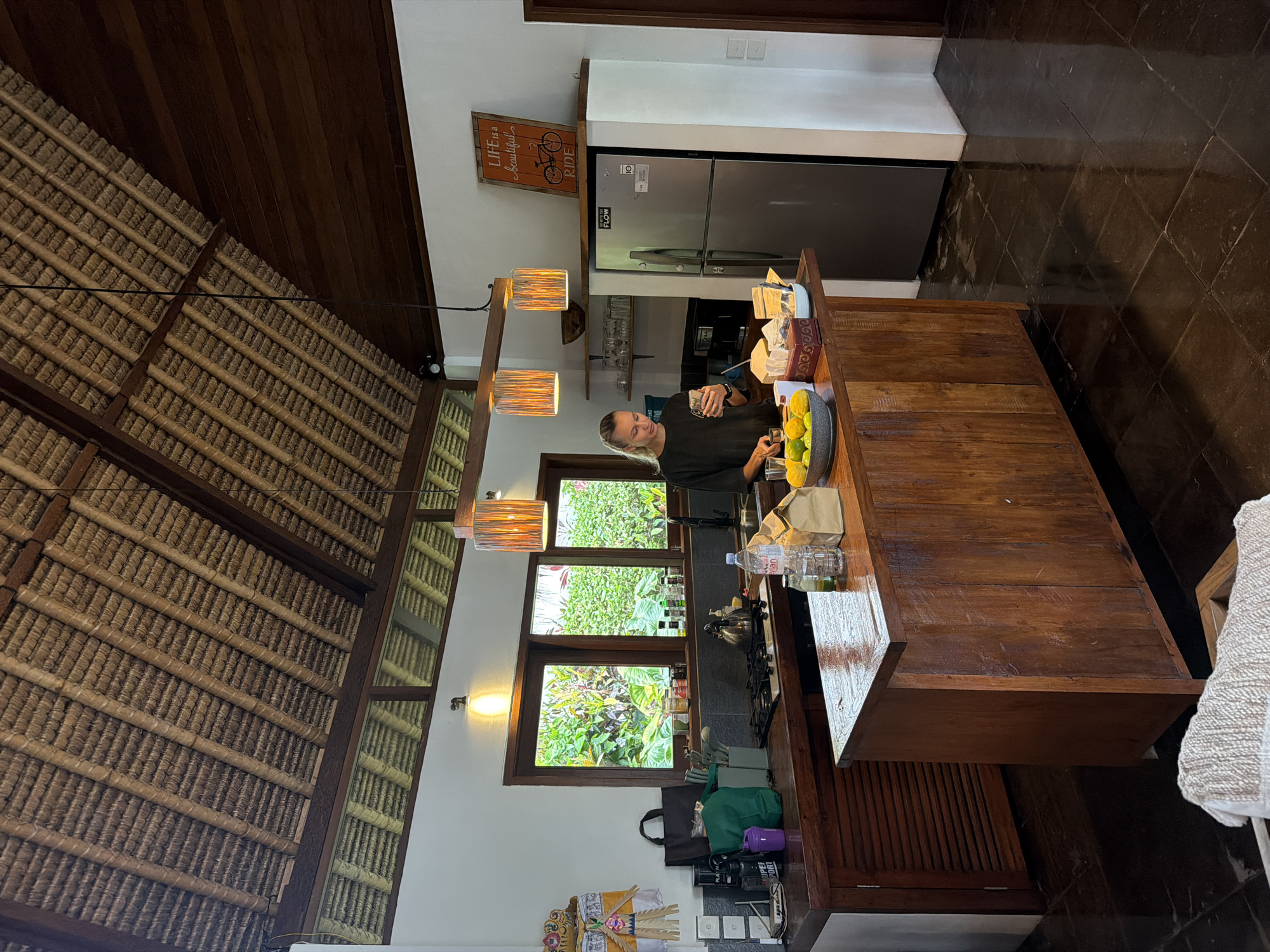 The image captures a bright, open-concept kitchen and living area, likely within a villa or home in a tropical setting such as Ubud District, Indonesia. A person, a woman with blonde hair wearing a dark top, stands at a large wooden kitchen island, holding a smartphone in one hand and a glass in the other, suggesting she is preparing a drink or snack.
The room features a high, vaulted ceiling composed of dark wooden beams and intricate woven or thatched panels. Several modern pendant lights with textured shades hang above the island, illuminating the space. Large windows behind the kitchen counter offer a view of lush green tropical foliage and plants, indicating daytime and an outdoor-oriented environment.
On the wooden kitchen island, a bowl of citrus fruits (likely limes or lemons), various bottles of water and other liquids, and a small coffee maker or kettle are visible. Behind the island, a stainless steel LG refrigerator is prominent, featuring an "LG" logo, a "10 YEAR WARRANTY" sticker, and a "FLOW" sticker. Above the refrigerator, a framed orange sign with a bicycle graphic reads: "LIFE is a beautiful RIDE".
The flooring is dark, possibly marble or stone tiles, and the overall aesthetic is a blend of rustic wooden elements with modern appliances and open design. On a lower counter to the left, a traditional Balinese offering (Canang Sari) is placed, a strong cultural cue for the Indonesian location. The scene conveys a relaxed, natural ambiance characteristic of a tropical resort or home.
