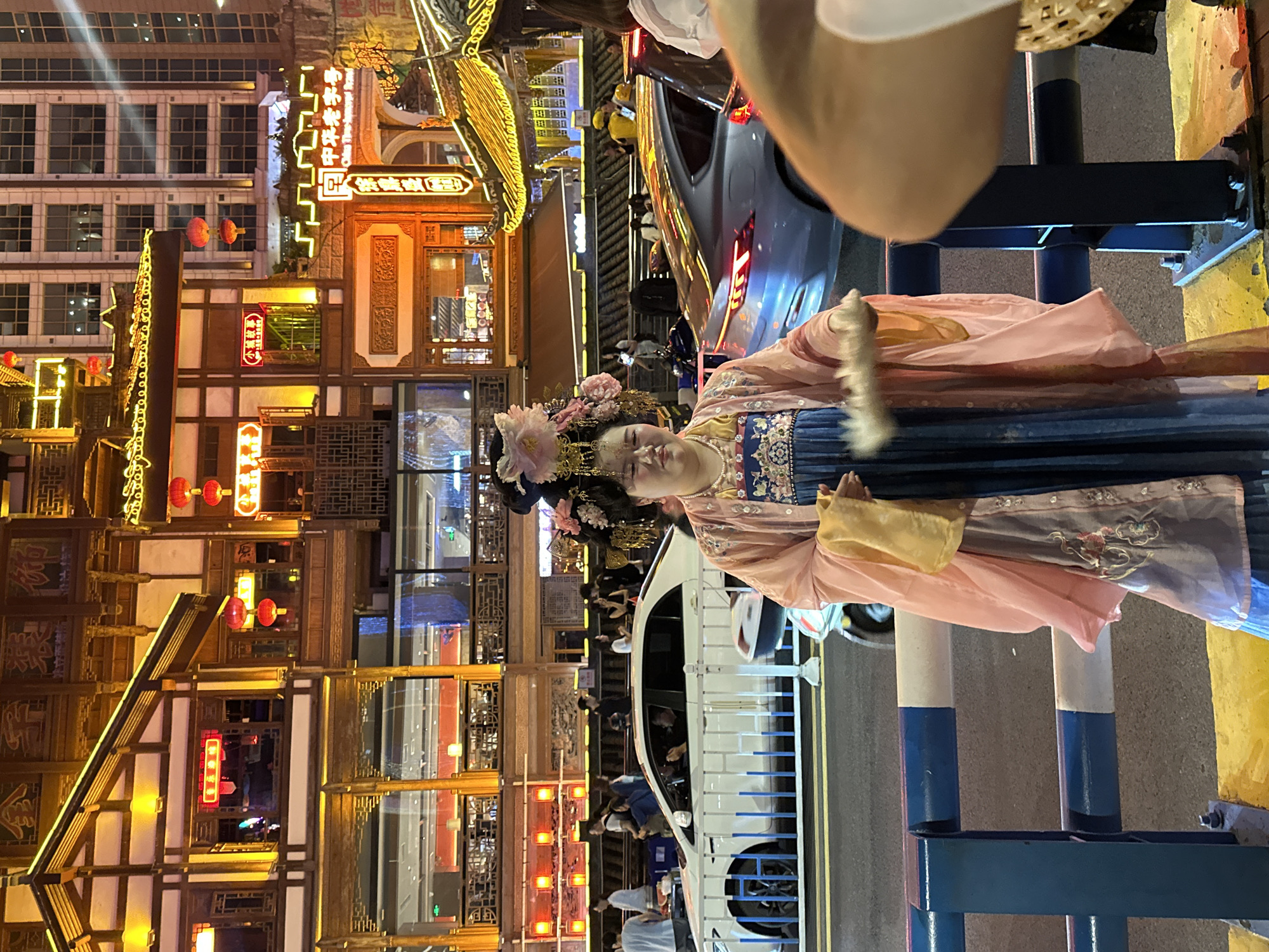 A street scene at night in the Yuzhong District of China, centered on a person dressed in a traditional Chinese garment standing by a car. The person is facing the camera and is adorned with a large, ornate floral headdress. Buildings with traditional Chinese architectural elements line the street in the background, with illuminated signs and lanterns. A white service truck is positioned on the street behind her as well. Pedestrians are also present. The scene includes street barriers in the foreground with cylindrical posts painted in blue and white.