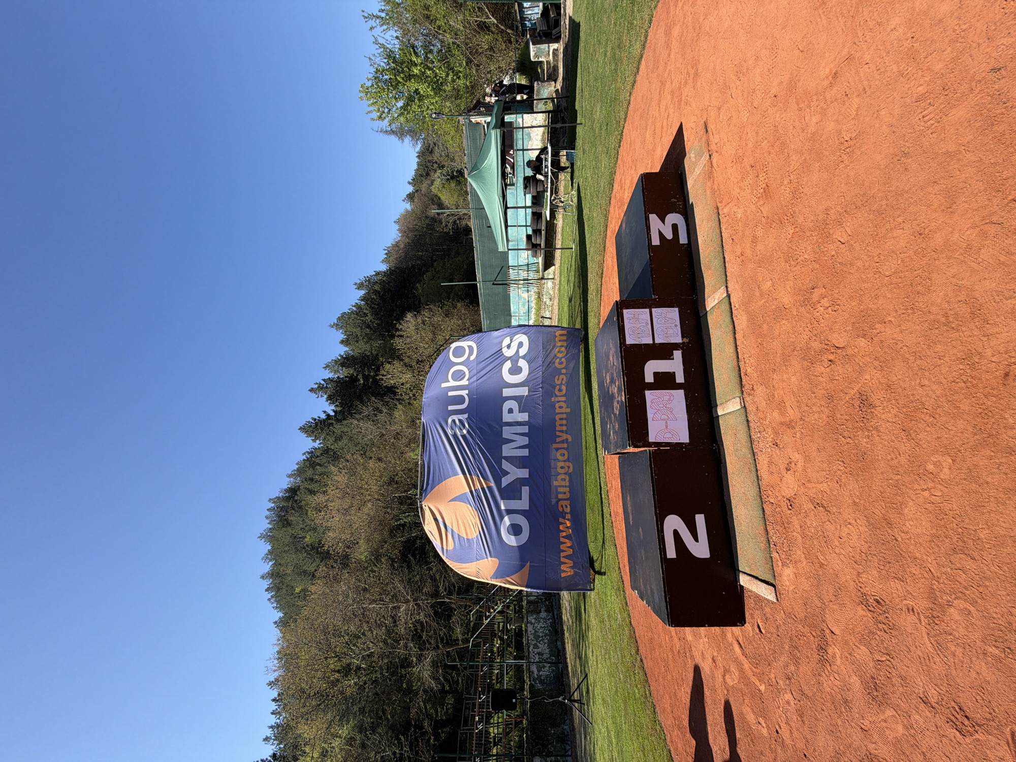 The image shows an outdoor scene, likely a sports event in Blagoevgrad, Bulgaria. A podium with "1", "2", and "3" marked on the front is present, positioned on a surface that appears to be a tennis court. A banner with the text "aubg OLYMPICS www.aubgolympics.com" stands near the podium. In the background, trees and possibly a sports facility are visible under a clear blue sky. The scene seems to be set up for an awards ceremony.