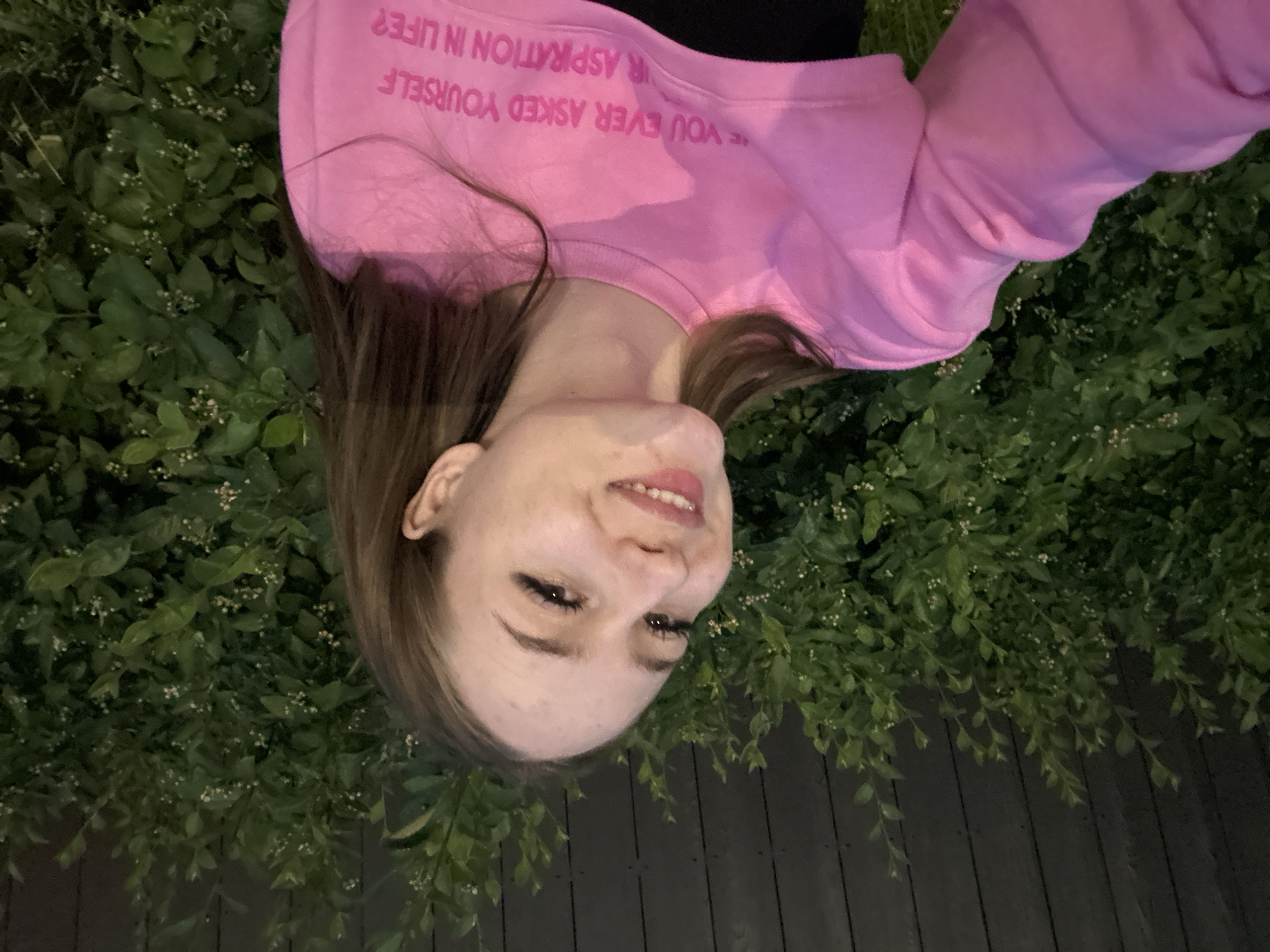 A young woman with long brown hair, wearing a pink cropped sweatshirt over a black top, is captured in a selfie outdoors. She is smiling, with her head tilted slightly to the left. Behind her, a dark wooden fence is partially visible, overlaid with lush green foliage. The lighting suggests it is evening or dusk. The text on her sweatshirt reads, "YOU EVER ASKED YOURSELF YOUR ASPIRATION IN LIFE?". The setting is an outdoor area, likely a backyard or garden, with no specific urban landmarks to identify a city.