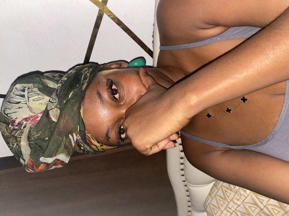 A dark-skinned woman wearing a floral headscarf is angled towards the camera, partially obscuring her face with her hand, featuring a red-painted fingernail. She dons false eyelashes and a green earbud. A gold chain necklace with clover-shaped embellishments drapes across her bare chest. She wears a gray thin-strapped top, seated in a white chair with a patterned cushion and gold-accented decor in the background.