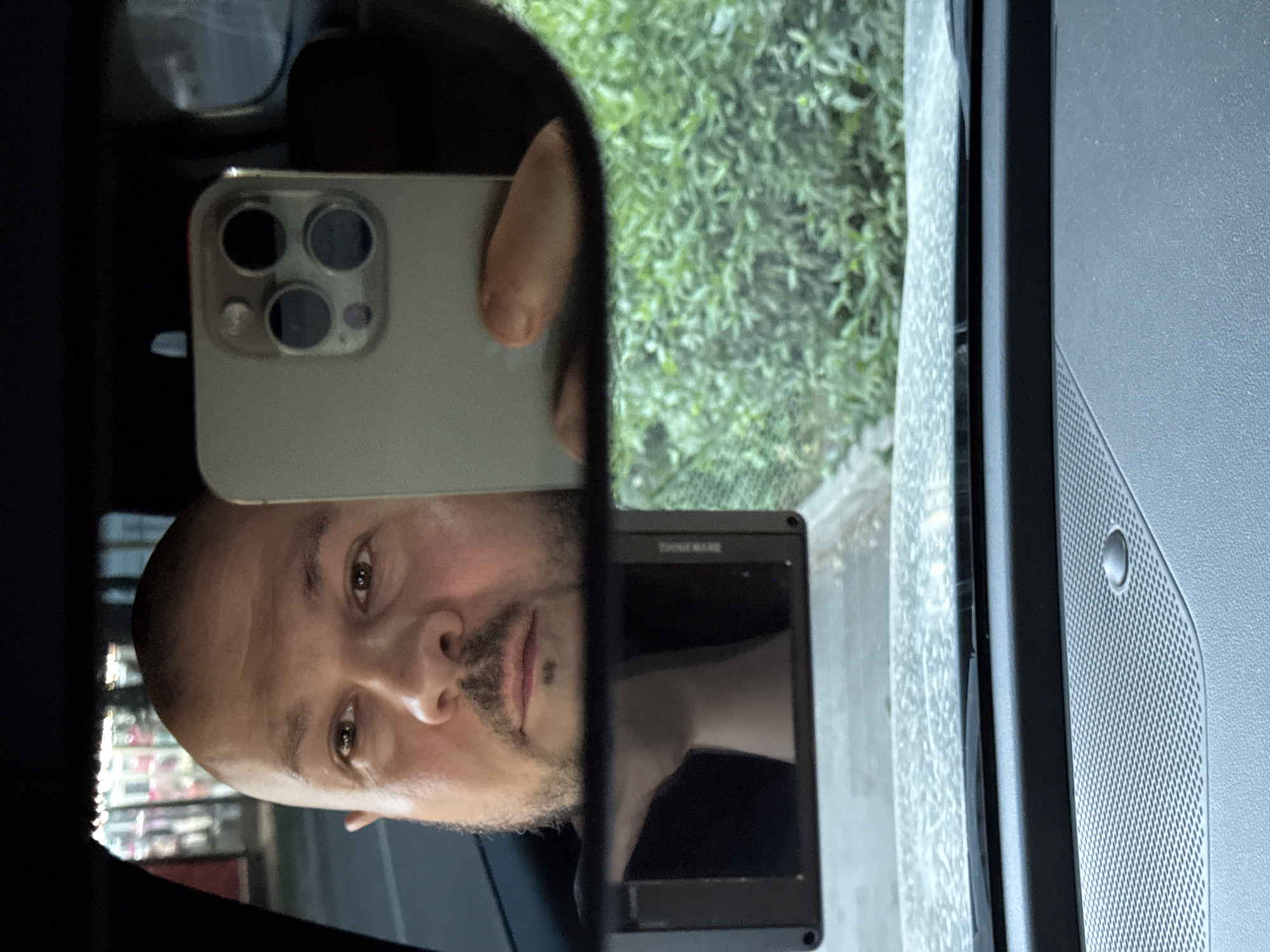 A person, identified as male by facial features and beard stubble, is reflected in a car's rearview mirror. The person is holding a smartphone, which is capturing the reflection. The phone is light gray with a prominent camera module featuring four circular lenses. The background seen in the mirror includes lush green foliage, suggesting an outdoor environment. In the foreground of the mirror, a black dashcam is mounted on the car's windshield, its screen displaying a reflection of the person's neck and shoulder. The overall scene is viewed from inside the car, looking towards the dashboard and windshield. The city of Almaty, Kazakhstan is not visually represented in the image.