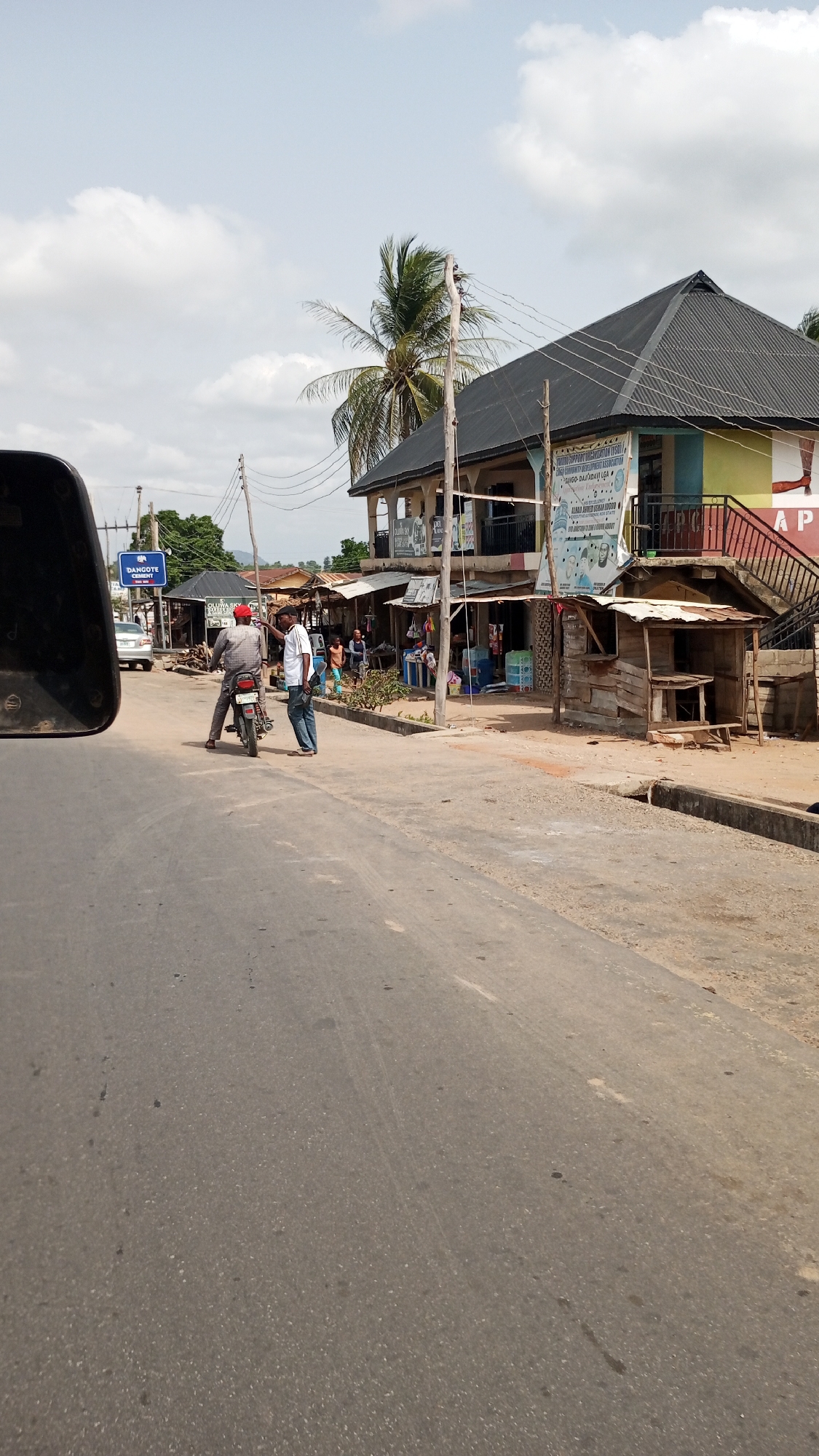 The image shows a street scene in Zango-ndaji, Nigeria. The road is asphalt and stretches into the distance. On the right side of the road are several buildings, including a two-story structure with a dark roof and a yellow and blue facade. A sign hangs on the front of the building. There is a small wooden structure in front of it, with a roof. On the left side of the road are other buildings. A sign on a pole says "DANGOTE CEMENT". In the middle of the road, two men stand next to a parked motorcycle. One man is seated on the motorcycle, and the other man is standing next to him, appearing to be talking. A car is visible on the left side of the road in the distance. A palm tree and power lines are visible in the background. The sky is partly cloudy.