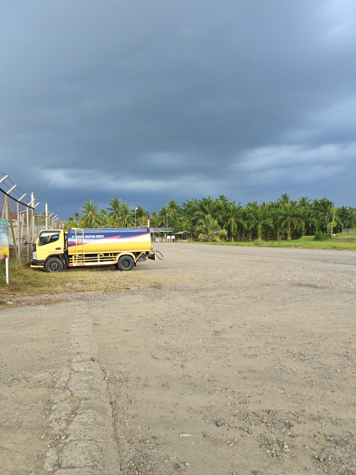 A yellow water truck with a blue and yellow tank is parked on a gravel lot next to a chain-link fence. The truck has the text "PT. BERRA PRATION GROUP" on its tank. In the background, there is a row of palm trees and a cloudy sky. The scene appears to be in Gampongpaya, Indonesia.