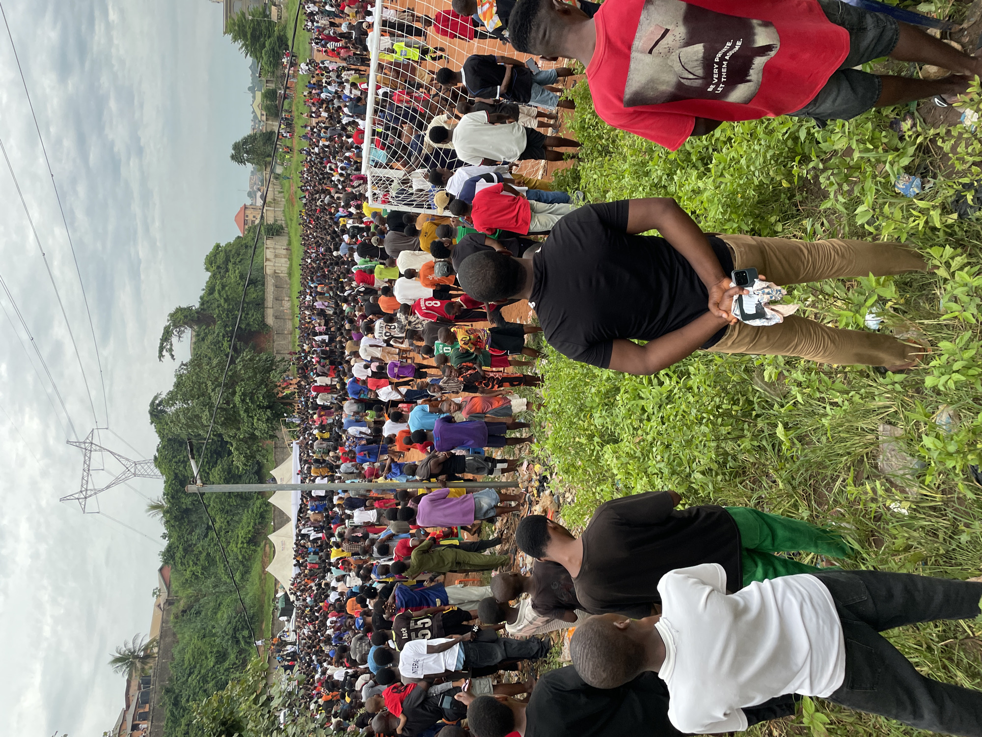An outdoor, wide-angle, eye-level shot captures a large gathering of people, many of whom are standing close together. The crowd is dispersed across a grassy area, with a soccer field and goal visible in the mid-ground. Tents are set up in the background, and beyond that, buildings and lush green hills with some trees are visible. A prominent metal tower with electrical lines extends upwards into the overcast sky. In the foreground, four individuals are partially visible from behind, facing away from the camera towards the crowd. The ground is uneven and covered with dense green vegetation, with some litter scattered throughout. The overall atmosphere appears to be that of an outdoor event or gathering.