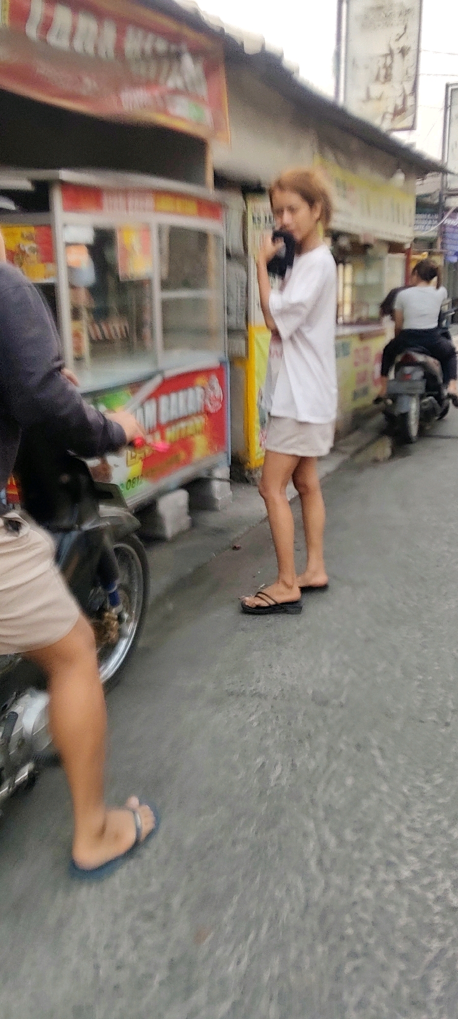 In Mekarmukti, Indonesia, a person with light brown hair stands on a street, holding a black object to their ear, suggesting a phone call. They are wearing a loose white t-shirt and a light-colored skirt. The person is also wearing black flip-flops. Behind them, a scooter is parked, and another person is visible riding a scooter further down the street. To the left, a food cart with red and yellow signage is present. The street appears to be paved, and the overall environment suggests an outdoor urban setting.