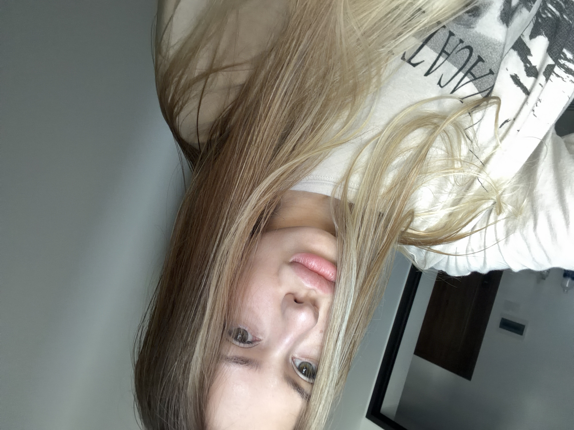 A young woman with long blonde hair is taking a selfie in what appears to be a room with light grey walls. She is wearing a white t-shirt with black text and graphics. The room is minimally furnished, with a dark door visible in the background on the left. The lighting suggests it is daytime. The woman's facial features are prominent in the close-up shot, with her lips slightly parted and her eyes looking towards the camera. There is no indication of the city of Đà Nẵng, Vietnam in the image.