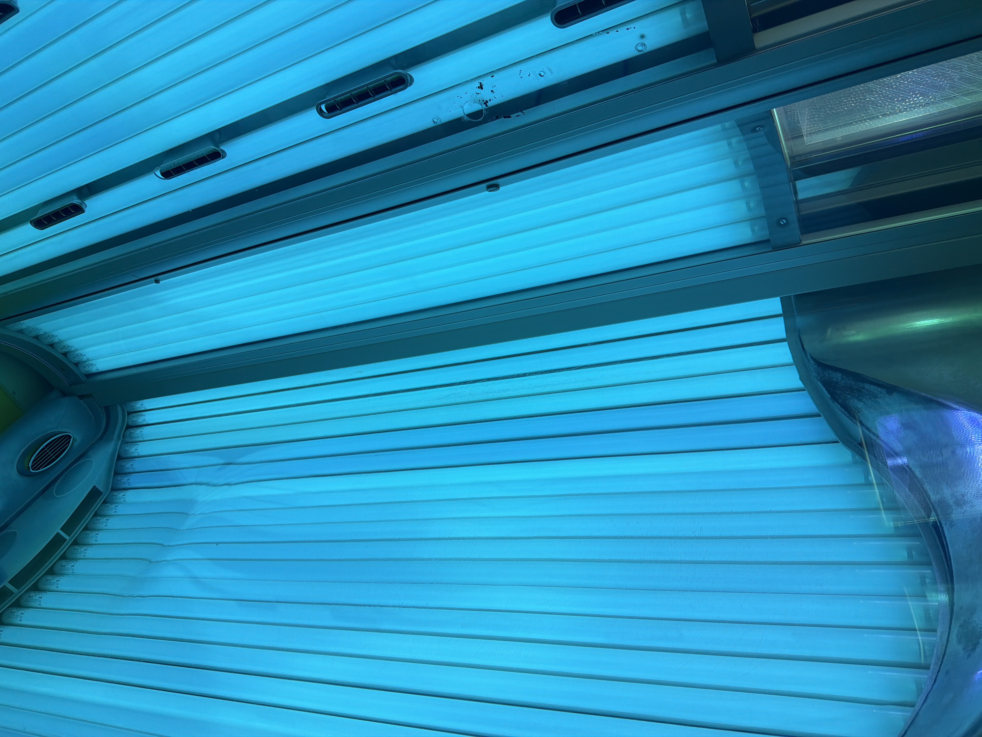 The image shows the interior of a tanning bed. The bed is illuminated by blue UV lights, which are arranged vertically along the walls and ceiling. The surfaces are mostly smooth and reflective, with some metallic accents and vents visible. The overall impression is one of artificial lighting and a confined space designed for tanning. The location in Kyiv, Ukraine is not determinable from the image itself.