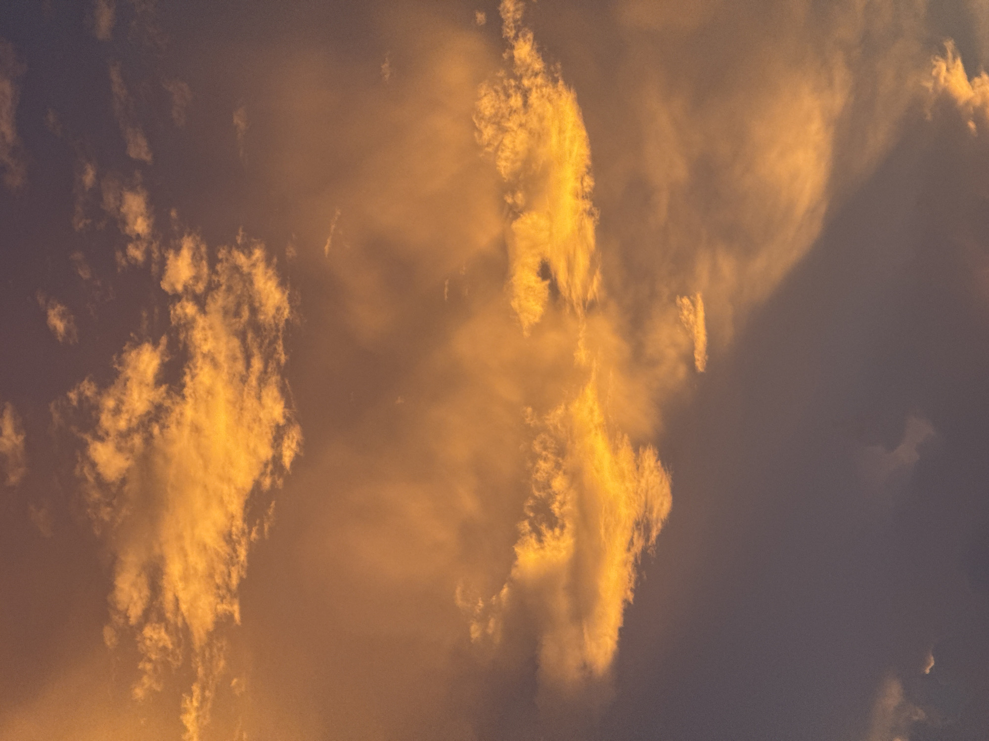 The image depicts a sky filled with clouds illuminated by the warm hues of sunset or sunrise. The clouds are predominantly orange and yellow, with some darker, purplish areas suggesting shadows or the approaching twilight. The overall atmosphere is dramatic and colorful, characteristic of atmospheric light phenomena. There are no discernible people, subjects, objects, or actions visible in this image, which solely captures the sky and its cloud formations. The location is specified as Goviyapana, Sri Lanka, suggesting this is a natural landscape scene.