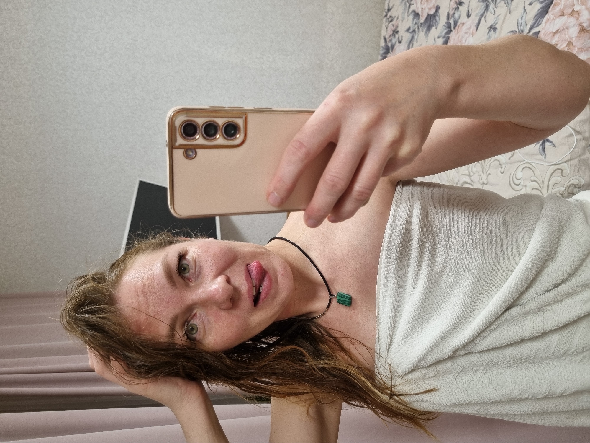 An indoor selfie shows a woman with a playful expression, her head tilted, sticking out her tongue towards the camera. She has light brown, possibly damp, hair, green eyes, and visible freckles on her face. Around her neck, she wears a black cord necklace with a rectangular green pendant, likely malachite. She is wrapped in a white, textured towel, suggesting she may have recently showered.
She holds a light gold or beige smartphone, possibly a Samsung Galaxy model, which features three camera lenses on its back, indicating she is using the front-facing camera.
The background reveals a home setting. To her left (viewer's right in an upright orientation), there is a wall with subtle light-colored patterned wallpaper. To her right (viewer's left), a bed is visible with a floral patterned duvet or sheet, featuring pink and grey-blue botanical designs on a light background. A plain light brown fabric, possibly a curtain or additional bedding, is present in the foreground. A partial view of a black object, resembling a laptop or tablet, is seen behind the smartphone. The scene conveys a casual and relaxed atmosphere. No text is visible in the image.