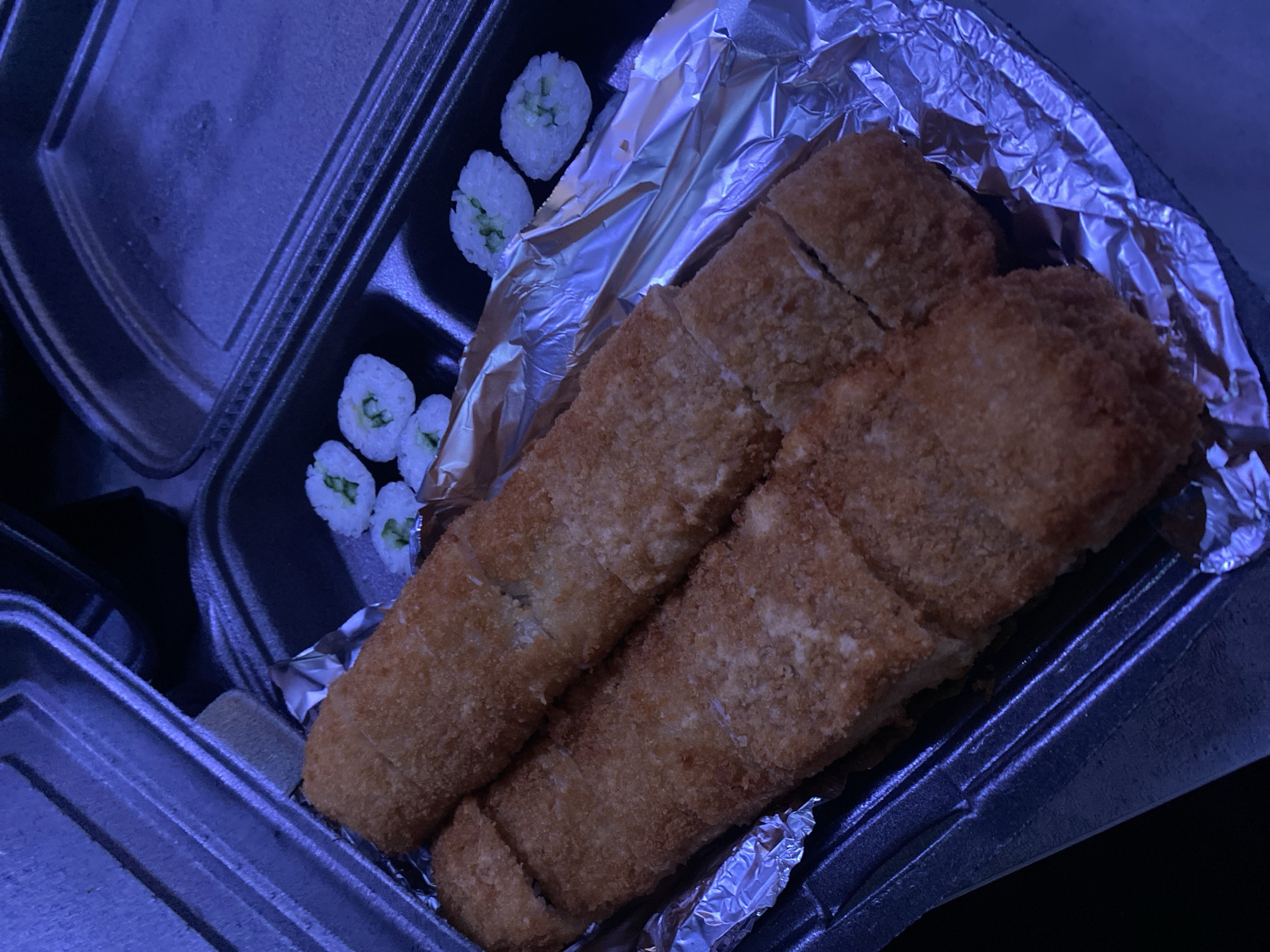 The image shows a take-out container of food, likely sushi. The container has two compartments. One compartment holds several pieces of sushi rolls filled with a green substance, possibly cucumber. The other compartment contains fried food, possibly tonkatsu or katsu, wrapped in aluminum foil. The setting appears to be indoors due to the blue lighting. There is no information about the city of Izhevsk, Russia or any people in the image.