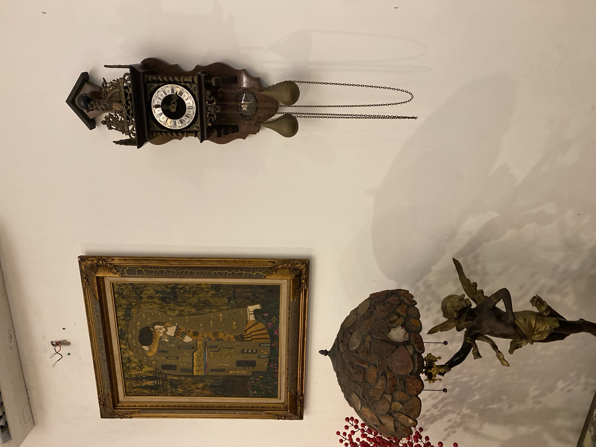 The image captures an interior wall adorned with decorative items. Dominating the upper left is a framed painting, depicting two figures embracing, rendered in a style reminiscent of Gustav Klimt's "The Kiss." The painting is set within an ornate gold frame. To the right, a traditional pendulum clock hangs on the wall. This clock features a dark wooden casing, a round face with Roman numerals, and two weights suspended by chains. Below the painting, a lamp with a mosaic-style shade rests on a stand shaped like a cherub. The cherub figure is bronze with gold accents, holding a decorative element above its head. A branch with red berries is visible to the left of the lamp. The wall itself is painted a neutral color. There is no visible indication of the city of Da Nang, Vietnam, in the image. The scene appears to be a still life arrangement in a domestic or gallery setting.