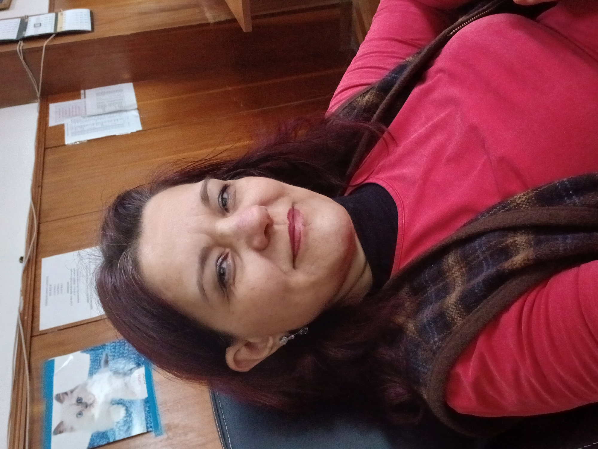 A woman is seen lying down on a black surface. She has dark reddish-brown hair, fair skin, and is wearing a red long-sleeved shirt with a black undershirt and a brown plaid shawl or vest. She's wearing a small earring on her left ear. Behind her is a wooden wall with a calendar and some papers attached. At the bottom left corner of the image, there's a picture of a white cat. The image does not provide information about the location or Vratsa, Bulgaria.