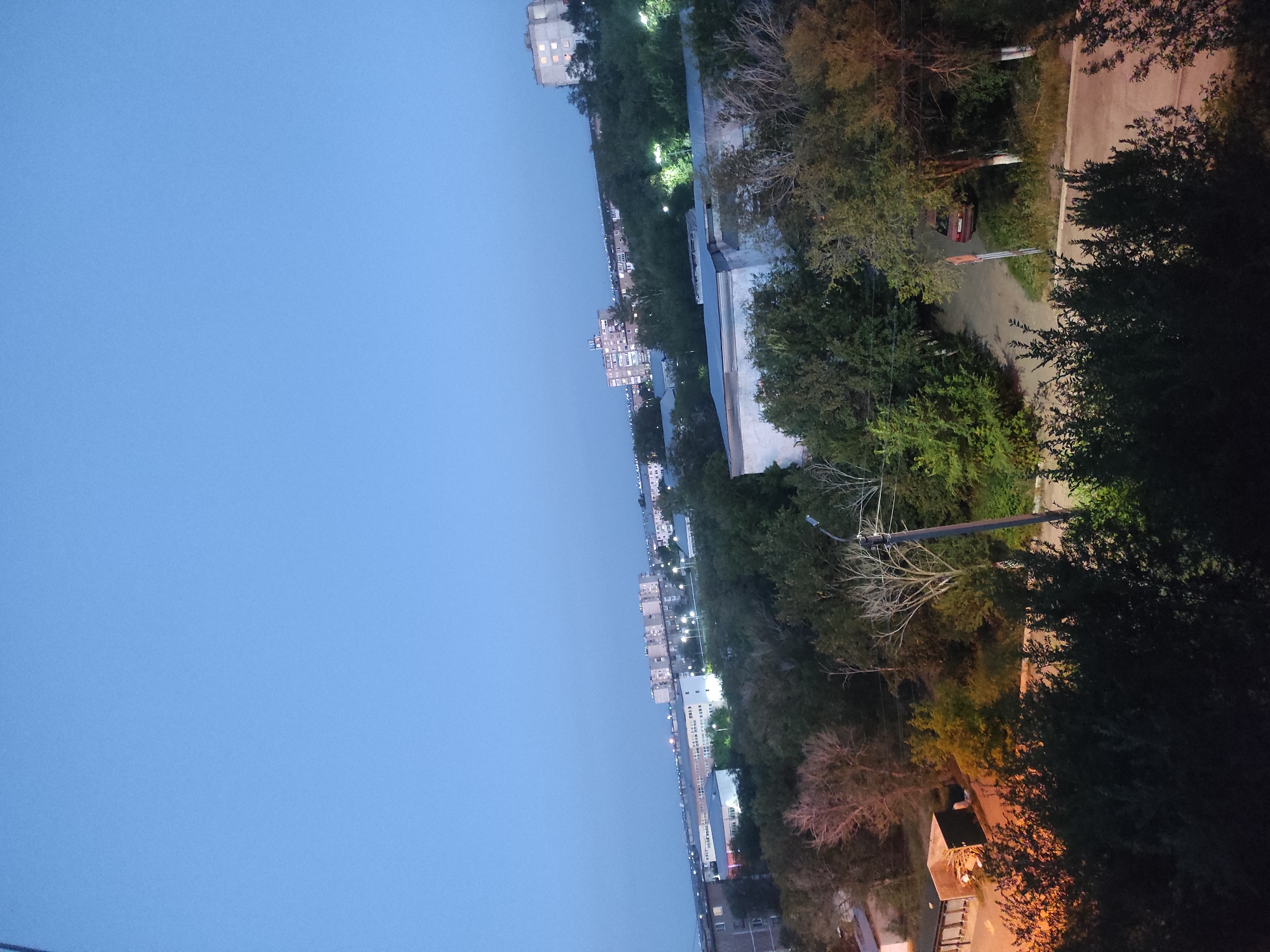 This is an aerial, high-angle shot of a residential area in Shakhtinsk, Kazakhstan, taken at dusk or dawn. The sky is a pale blue, transitioning to darker hues near the horizon. In the distance, apartment buildings of varying heights, some with illuminated windows, are visible, suggesting the presence of a populated urban environment. Trees and greenery are abundant, forming a dense canopy in the middle ground and foreground, obscuring much of the ground level. A few scattered streetlights are illuminated, casting pools of light on the scene. A dirt road winds through the trees, with a red car parked on its side. The overall impression is one of a quiet, somewhat overgrown neighborhood at the edge of a city.