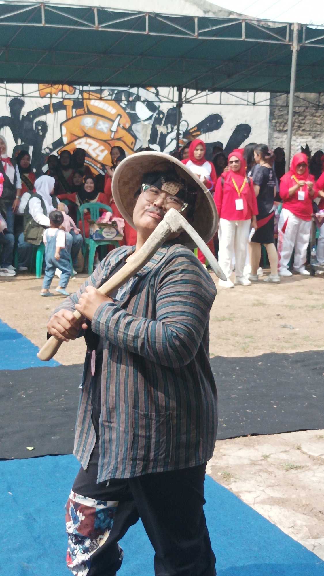 In Munjul, Indonesia, a person wearing a wide-brimmed hat and a striped shirt poses with a hoe. The individual has a mustache and glasses, and appears to be performing or participating in an event, given the crowd in the background. Many people in the background are wearing red outfits and hijabs. The setting seems to be an outdoor event with a green awning overhead and graffiti on a wall behind the crowd. The ground is covered with dirt and blue mats.