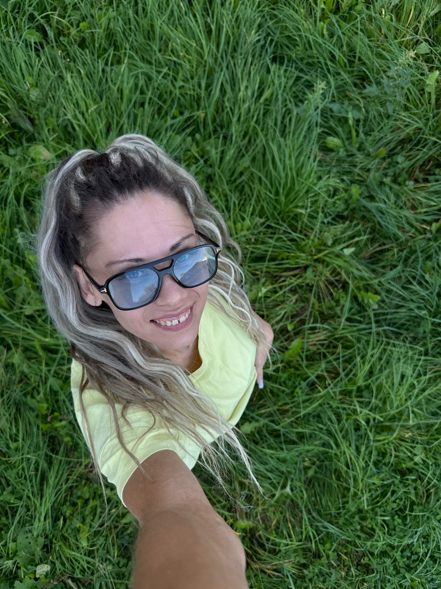 A woman with blonde hair in a high ponytail, wearing a yellow t-shirt and dark sunglasses, smiles at the camera while holding up her arm to take a selfie. She is lying on a grassy field, with the camera angled downwards. The field is covered in lush green grass and some small wildflowers. The scene appears to be outdoors, possibly in a park or a rural area. The specific location in the city of Rusanovo, Russia, cannot be determined from the image alone, as there are no identifying landmarks or context provided.