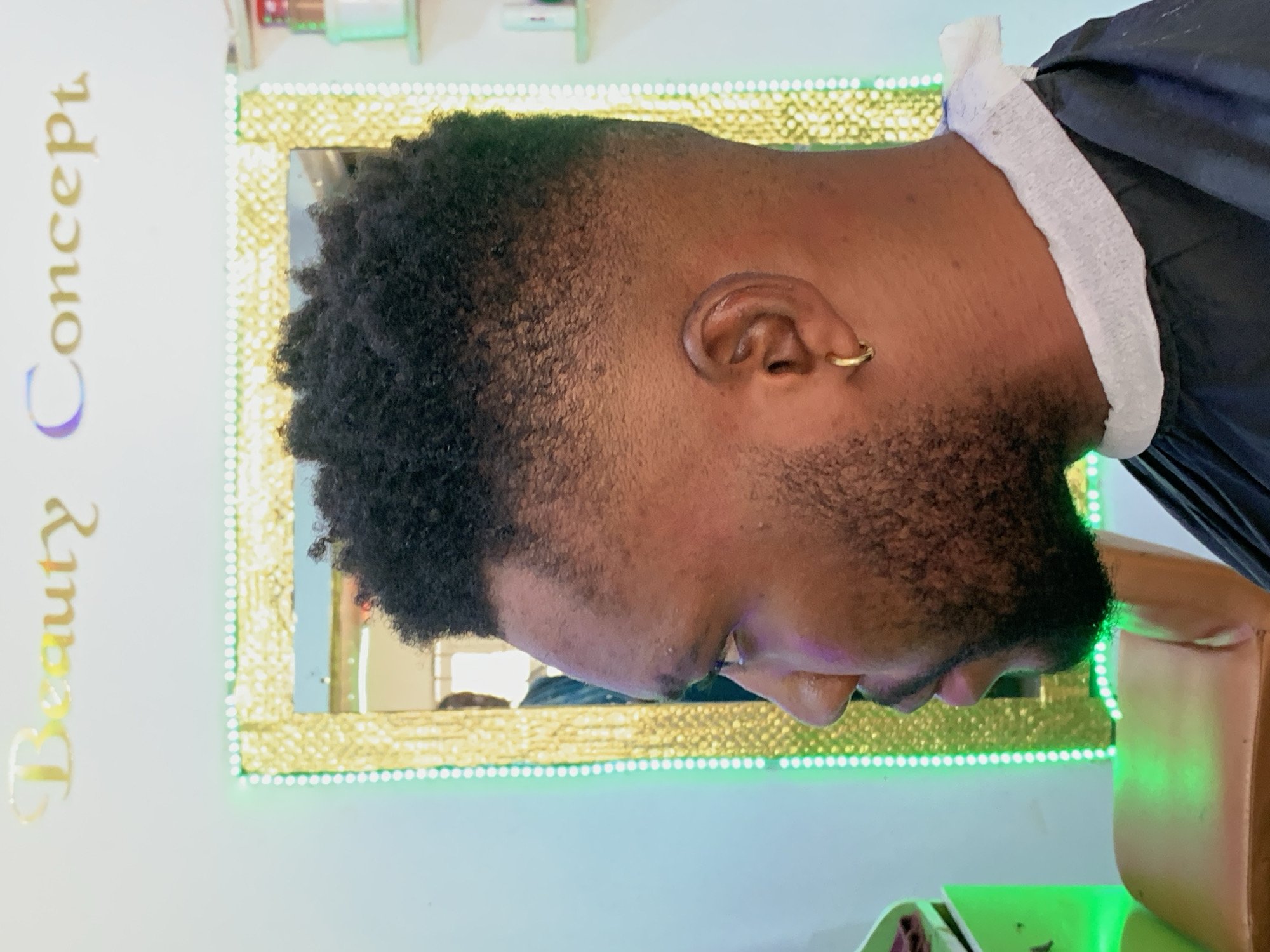 A medium shot depicts an individual wearing a black haircut apron with a white neck wrap. Their head is turned to the side, facing the left. The individual has short, dark, afro-textured hair, a beard, and a gold hoop earring in their left ear. The background includes a gold-framed mirror with green LED lights. To the left, "Beauty Concept" is displayed vertically in gold lettering on a light-colored wall.
It's not possible to confirm whether the image was taken in Ikorodu, Nigeria, based solely on the image.