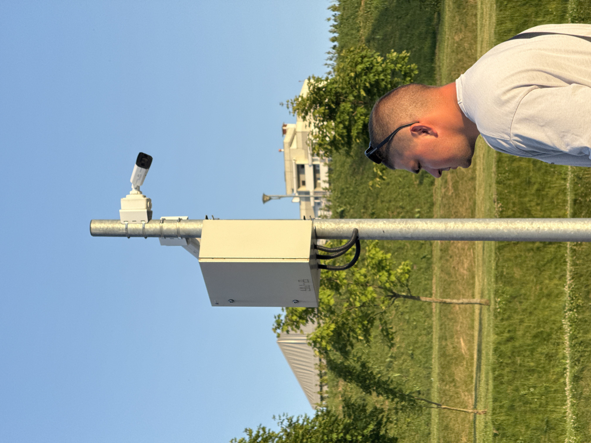 A medium shot captures a man from the shoulders up, his head tilted down as if observing something. He is wearing sunglasses and a light-colored t-shirt. The man is positioned on the right side of the frame, with his face partially obscured. In the background, a security camera is mounted on a pole, pointing upwards. The camera is white with a black lens. Adjacent to the camera is a light-colored box, likely housing equipment, with wires connected to it. The scene is outdoors, under a clear blue sky, suggesting it is daytime. Green grass covers the ground, with trees and a building visible in the middle ground. The overall setting appears to be an urban or suburban park area. There is no visible text in the image.