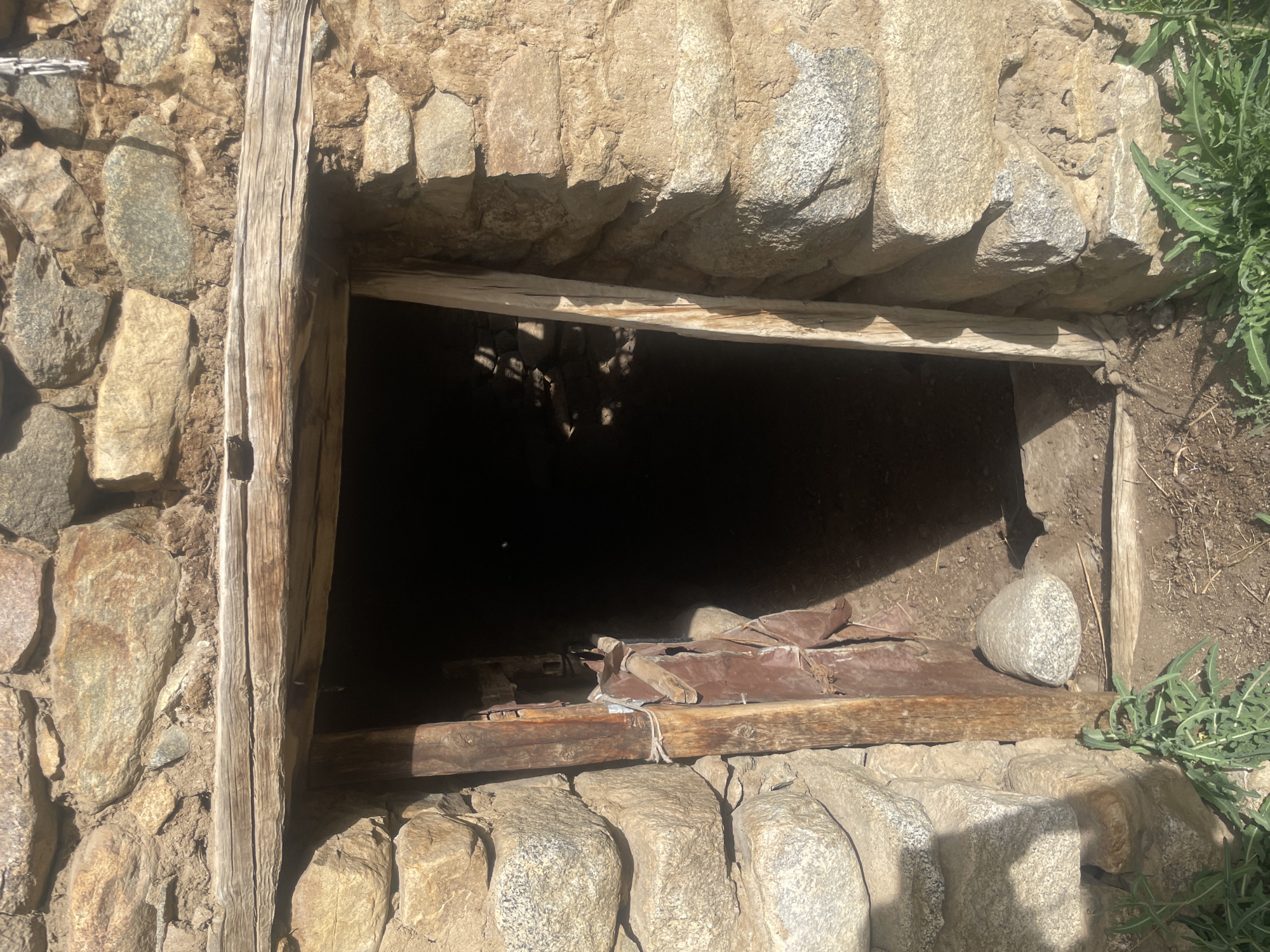 An outdoor, eye-level, medium shot shows the entrance to a dark, stone structure in Leh, India. The structure is built of irregular, tan and gray stones, held together with what appears to be mud mortar. A rough-hewn wooden beam spans the top of the entrance, and a narrow wooden post supports the left side of the doorway. The doorway itself is open, revealing a dark interior that is not discernible. Part of a reddish, weathered door is visible on the left side of the entrance, secured by twine to the wooden post. A few scattered rocks are visible on the ground at the entrance, and some green weeds grow in the foreground. The scene is lit by natural sunlight, casting shadows across the entrance and the surrounding stonework.