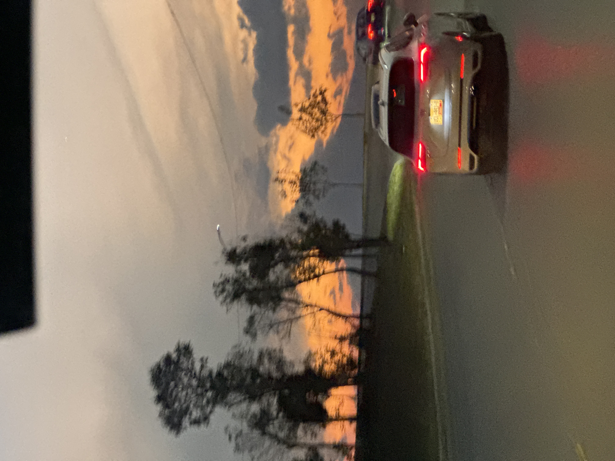 The image shows a dusk or dawn scene near a road in San Andrés Cholula, Mexico. The sky features layered clouds with orange hues suggestive of a sunrise or sunset. Silhouetted trees line the roadside. A white car with illuminated tail lights and a visible license plate is partially visible on the right side of the frame, as well as another vehicle in front of it. The scene is likely captured at night.