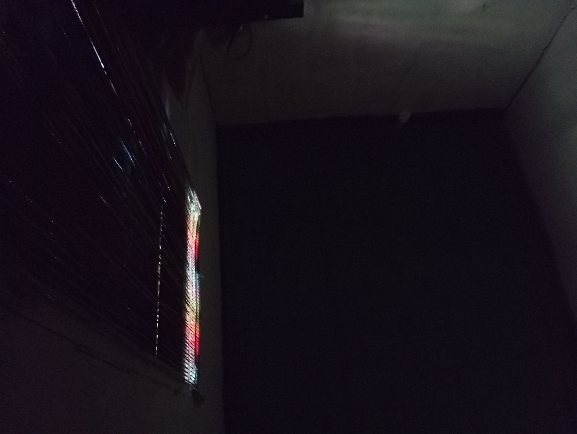 The image shows a dark interior scene with a faint light source on the right side, casting colorful reflections onto a vertical surface. The light appears to be coming from a string of lights with red, yellow, and white colors. The reflections are elongated and distorted, suggesting they are hitting a textured or uneven surface, possibly wrapped in plastic. The rest of the scene is predominantly dark, with hints of a wall on the left and a ceiling overhead. The overall impression is of a dimly lit space with an artificial light source creating abstract patterns. The location is specified as Rejosari, Indonesia, but there are no visual cues in the image to corroborate this detail. There are no people or distinct objects visible besides the implied light source and the reflective surface.