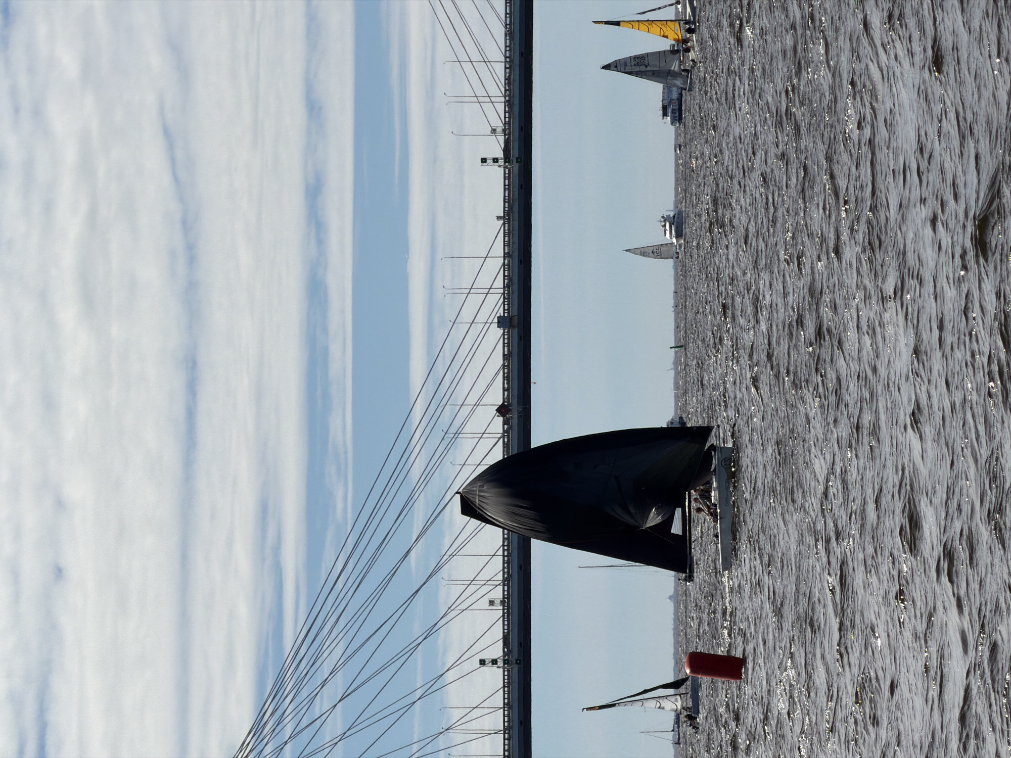 In Saint Petersburg, Russia, a fleet of sailboats navigates choppy, dark water under a partly cloudy sky. A large, modern cable-stayed bridge spans the horizon, its many cables stretching upwards. In the foreground, a black sail dominates the left side of the frame, obscuring a small white sailboat with a few people on board. To the left of this boat, a red cylindrical buoy floats on the water. Further in the distance, other sailboats with white and yellow sails are visible, along with larger vessels and a distant cityscape or landmass. The sun reflects off the water, creating bright highlights. The overall impression is of a sunny but breezy day on the water, with urban infrastructure as a backdrop.