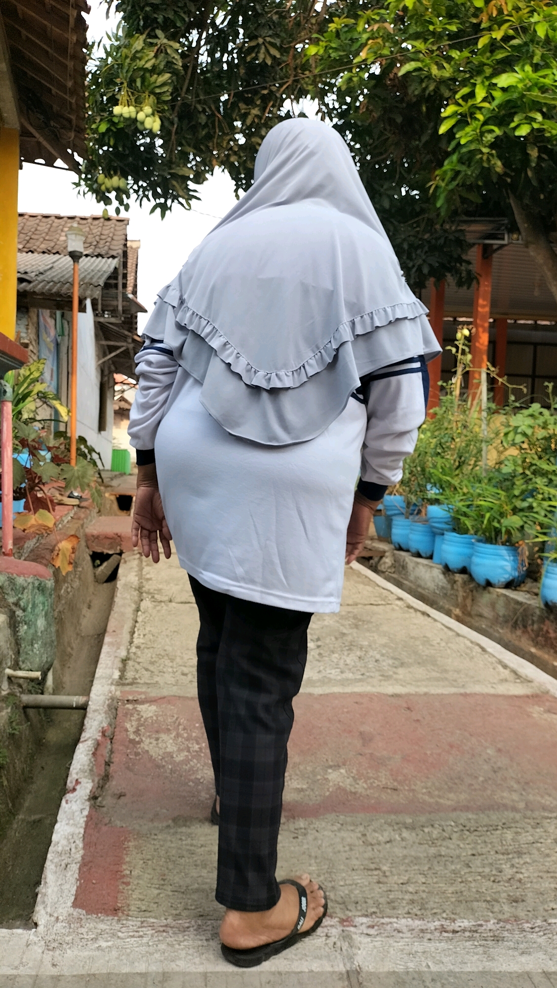 A person wearing a light gray hijab and a light gray tunic over dark plaid pants and black sandals walks away from the camera on a concrete walkway. The walkway is bordered by a shallow ditch on the left and plants in blue pots on the right. In the background, there is a small building with a yellow wall, a tiled roof, and a street lamp. Trees with green leaves and some unripe fruit are visible above the building. The scene appears to be in a residential area in Subah, Indonesia.