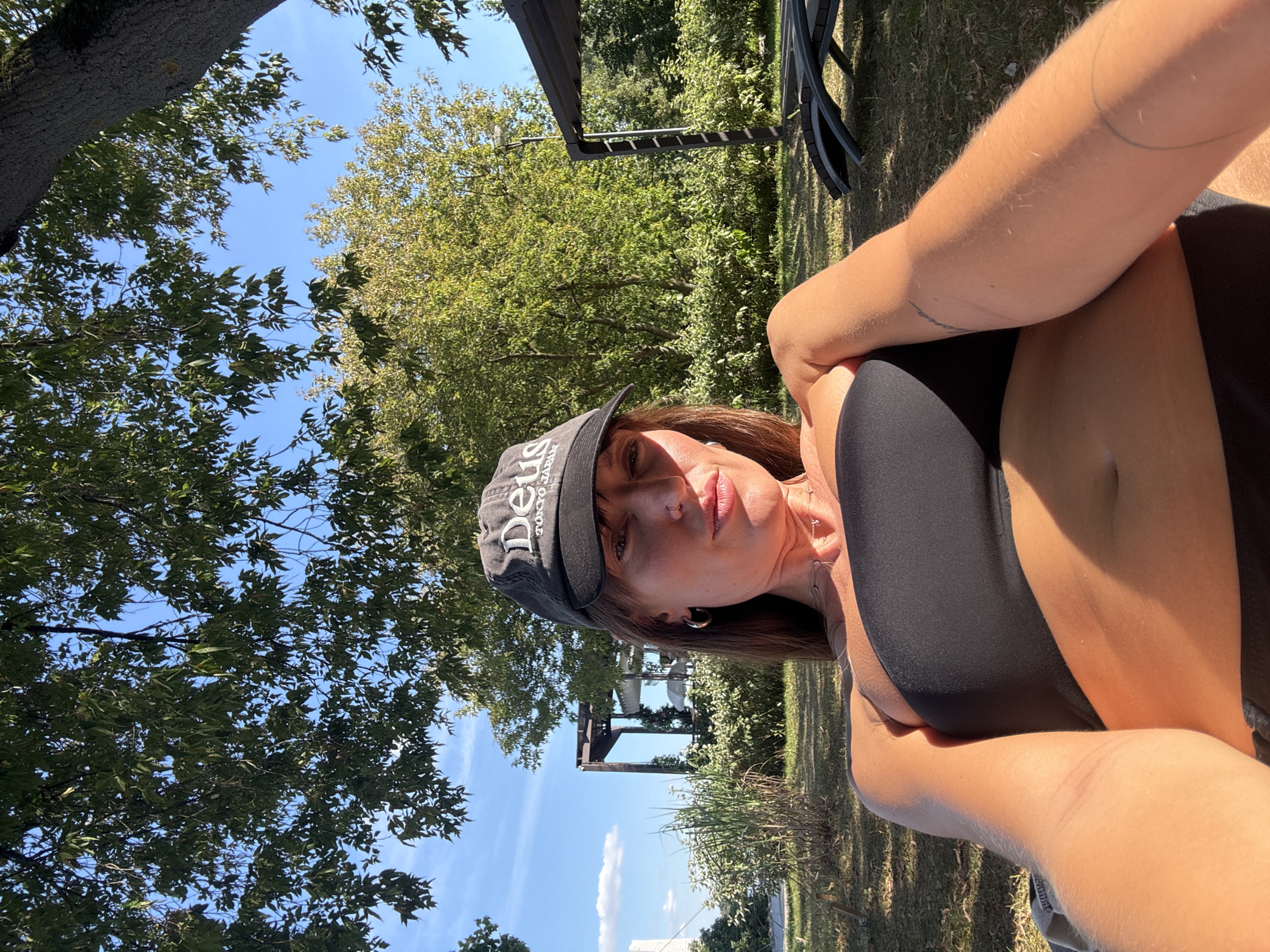 A woman wearing a black strapless top and a black baseball cap that says "Deus Tokyo Japan" is lying down outside in a grassy area. She has long brown hair, a nose ring, and hoop earrings. Her arms are visible, and there are tattoos on her left arm. In the background, there are trees, a pergola with white fabric, lounge chairs, and a bright blue sky with some clouds. The image appears to be taken on a sunny day.